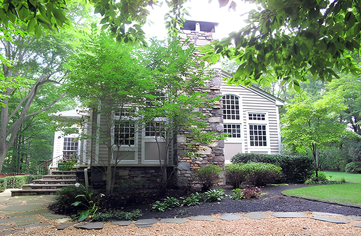 A house with a stone chimney is surrounded by trees and gravel