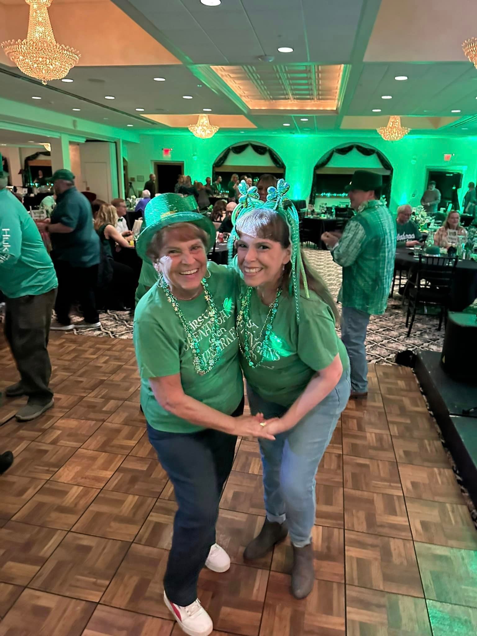 Two women are standing next to each other on a dance floor wearing green shirts and hats at a St. Patrick's Day Party.