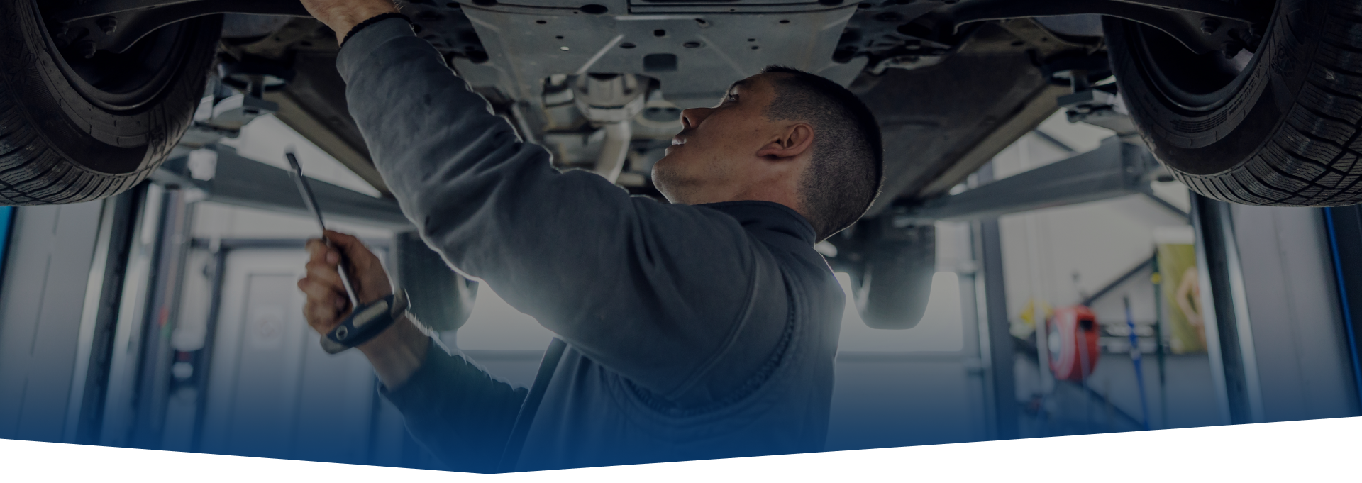 Mechanic working under a vehicle on a lift, inspecting the undercarriage with a tool