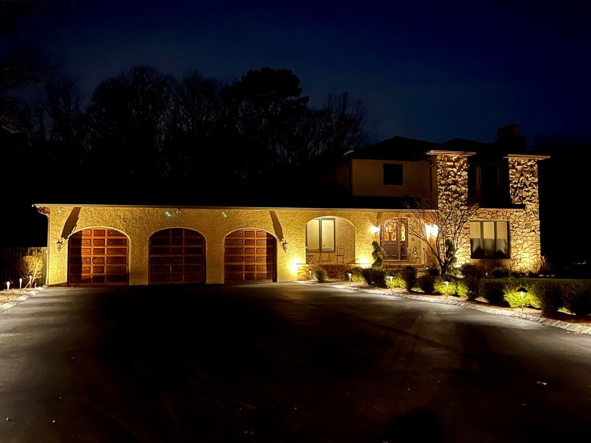 Night view of a house with illuminated driveway and facade. Brown garage doors, stone accents, and landscaping.