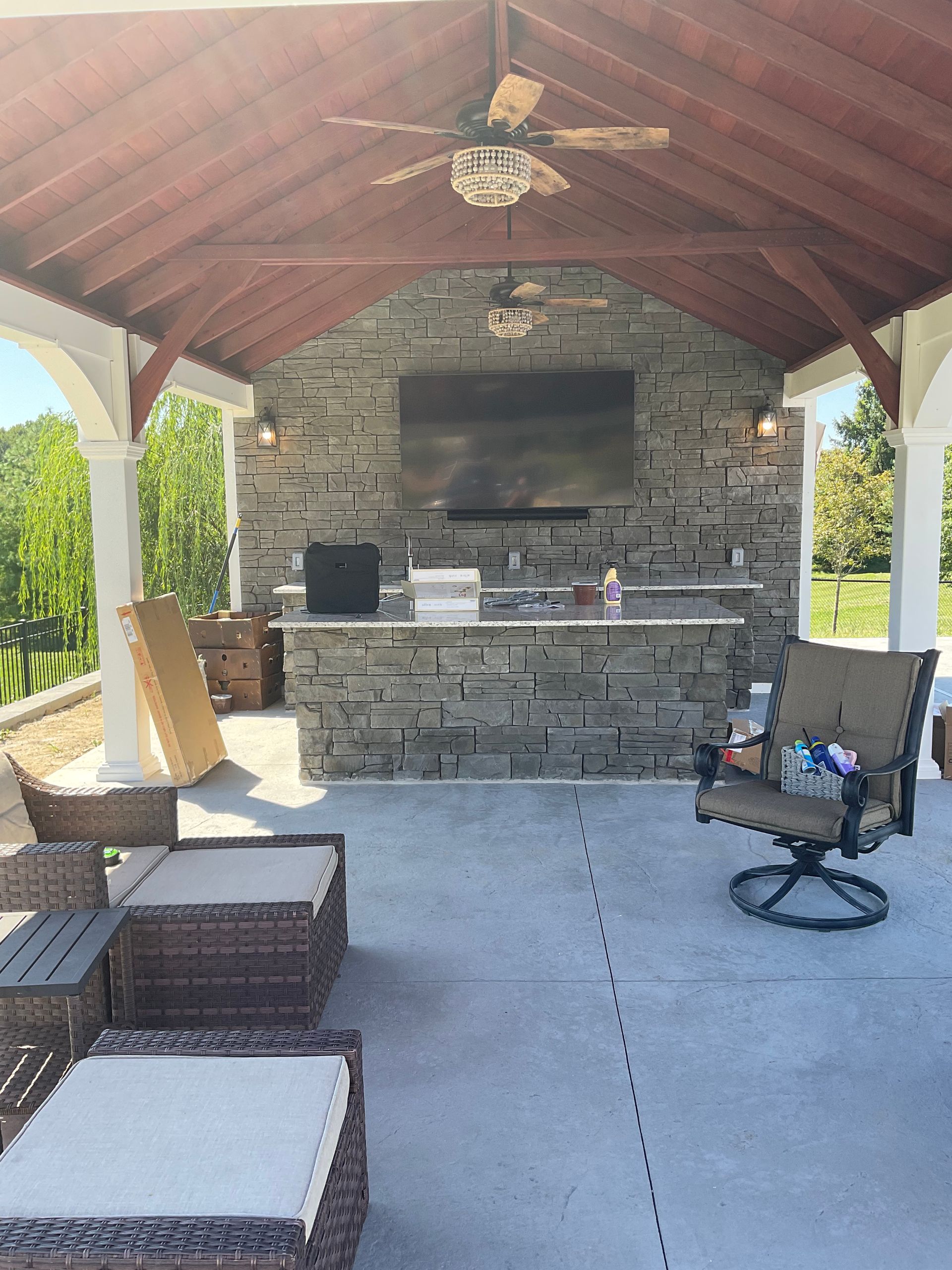 Outdoor kitchen under a covered patio with stone wall, TV, and seating.