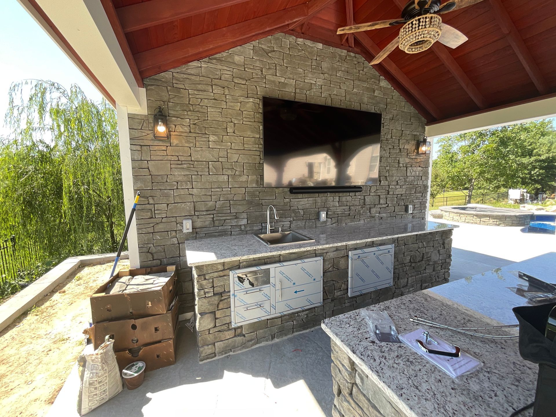 Outdoor kitchen with stone walls, a TV, sink, and cabinets under a roof.