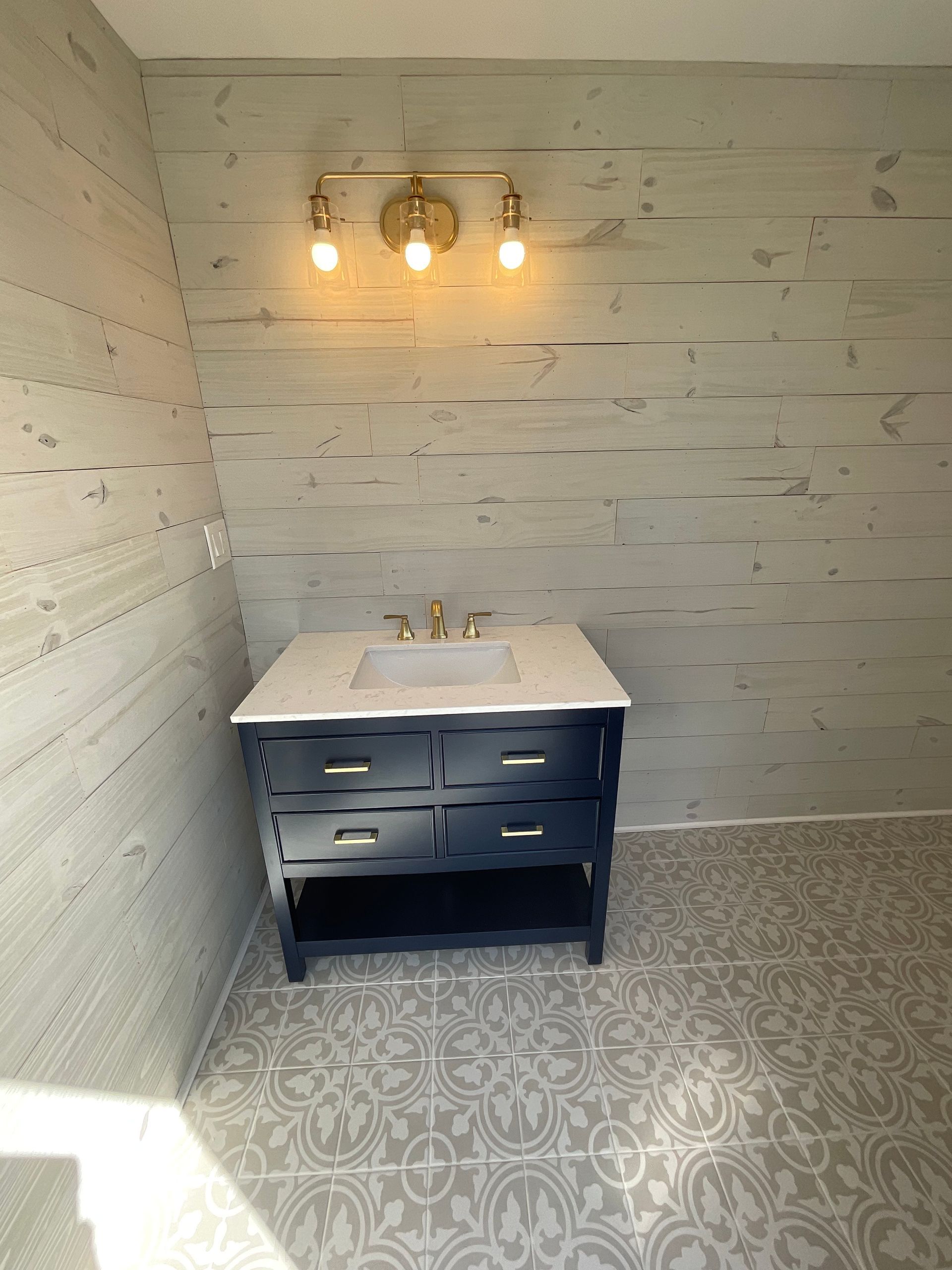 Bathroom with navy vanity, white countertop, gold fixtures, wood-look walls, and patterned tile floor.