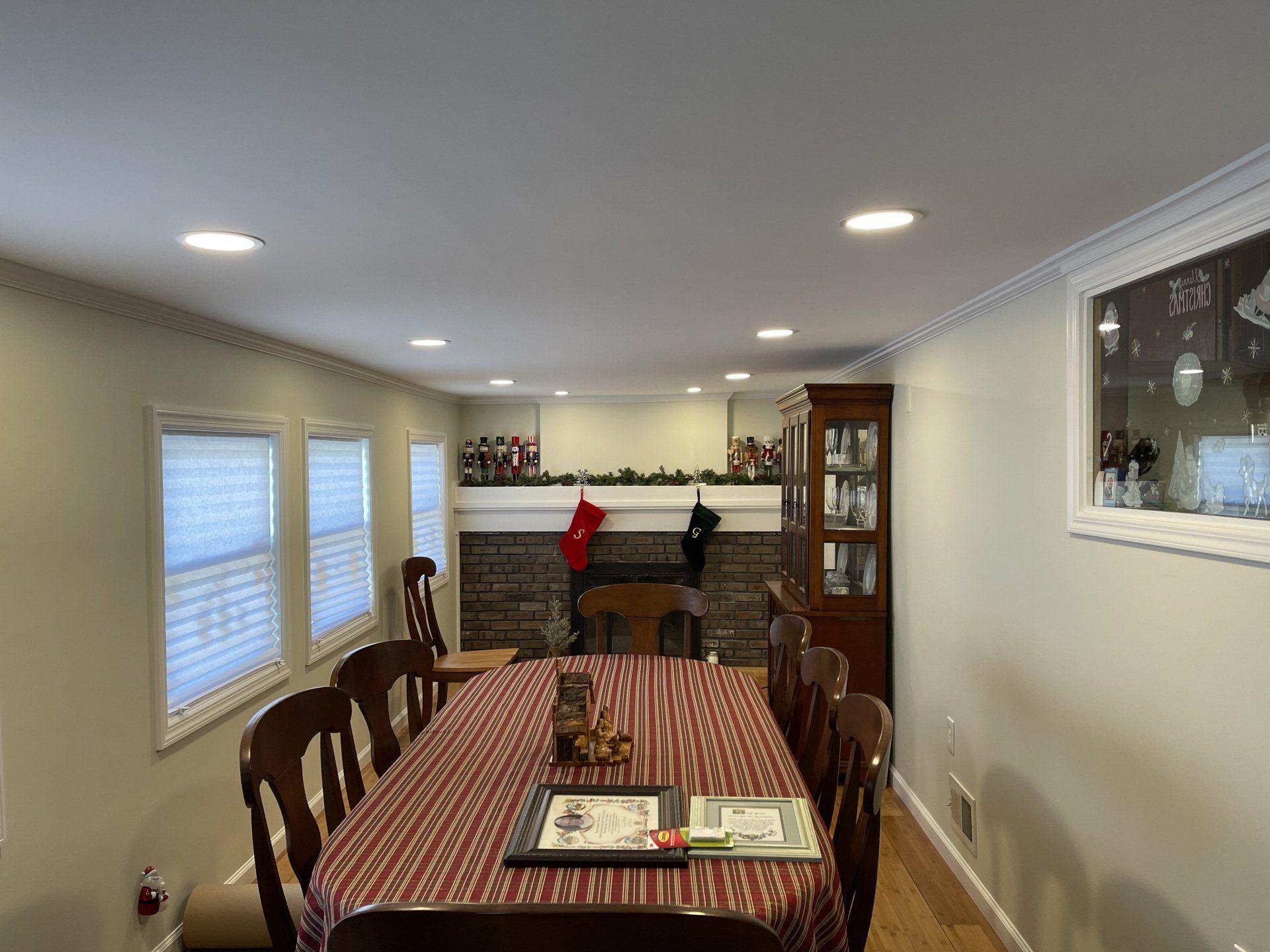 Dining room with table set for a meal, fireplace with stockings, wooden hutch, and windows.