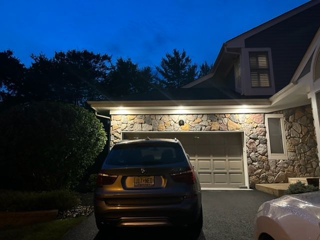 Dark car parked in front of a stone-faced garage, illuminated by overhead lights at dusk.