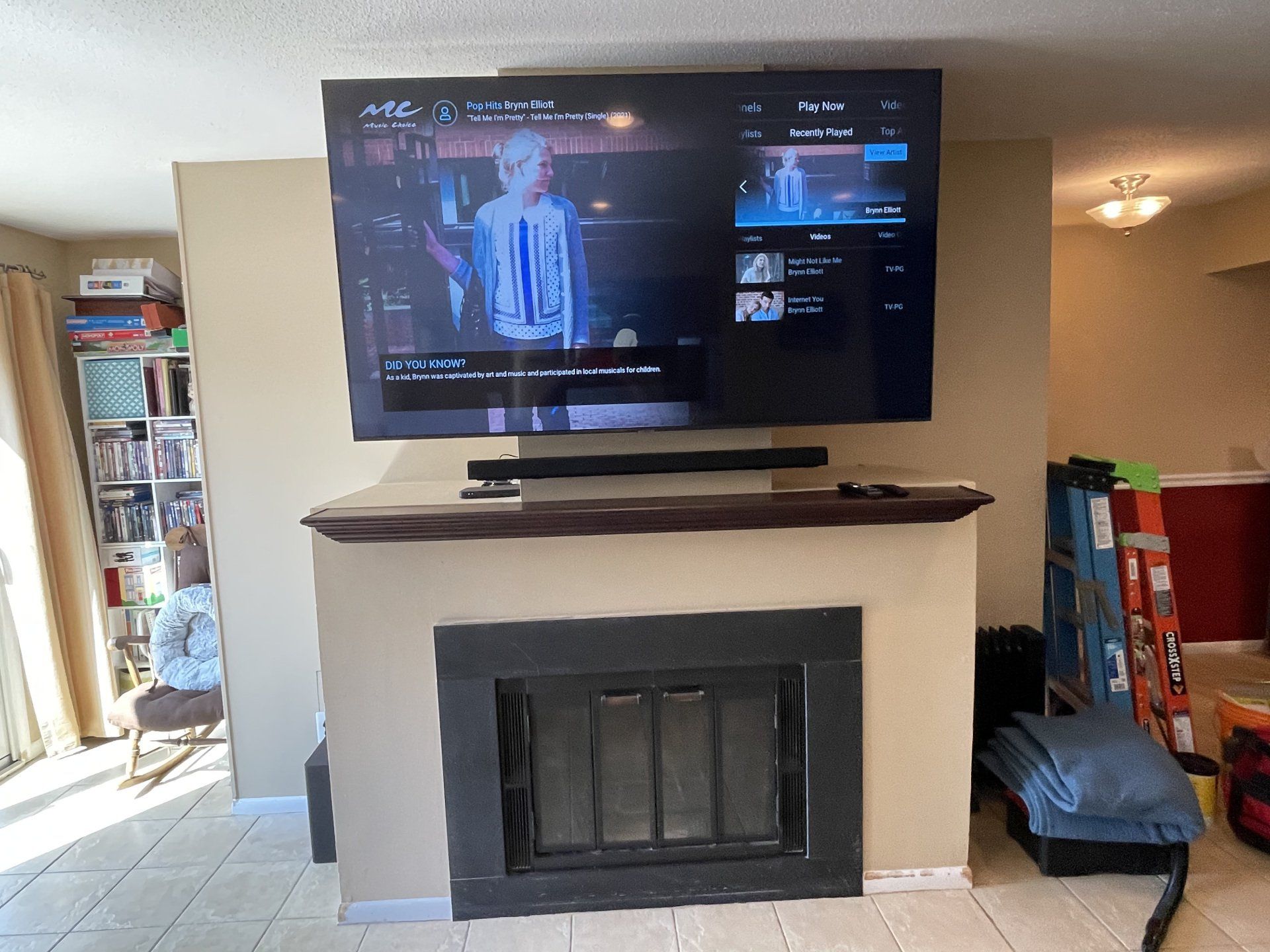 Large TV mounted above a fireplace, soundbar below it. Beige wall, tiled floor, room interior.
