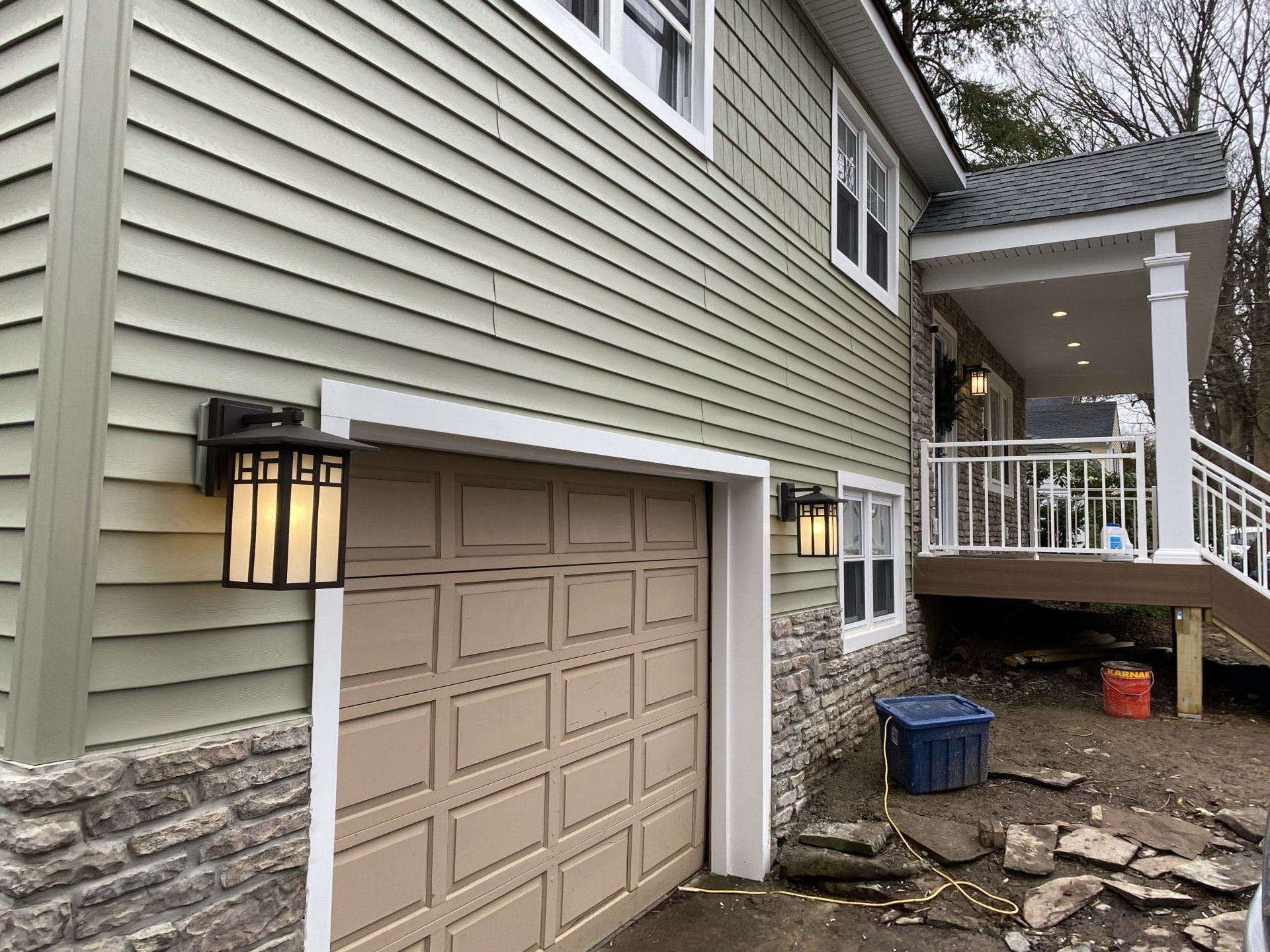 Garage and porch of a house with light green siding, tan garage door, and stone accents.