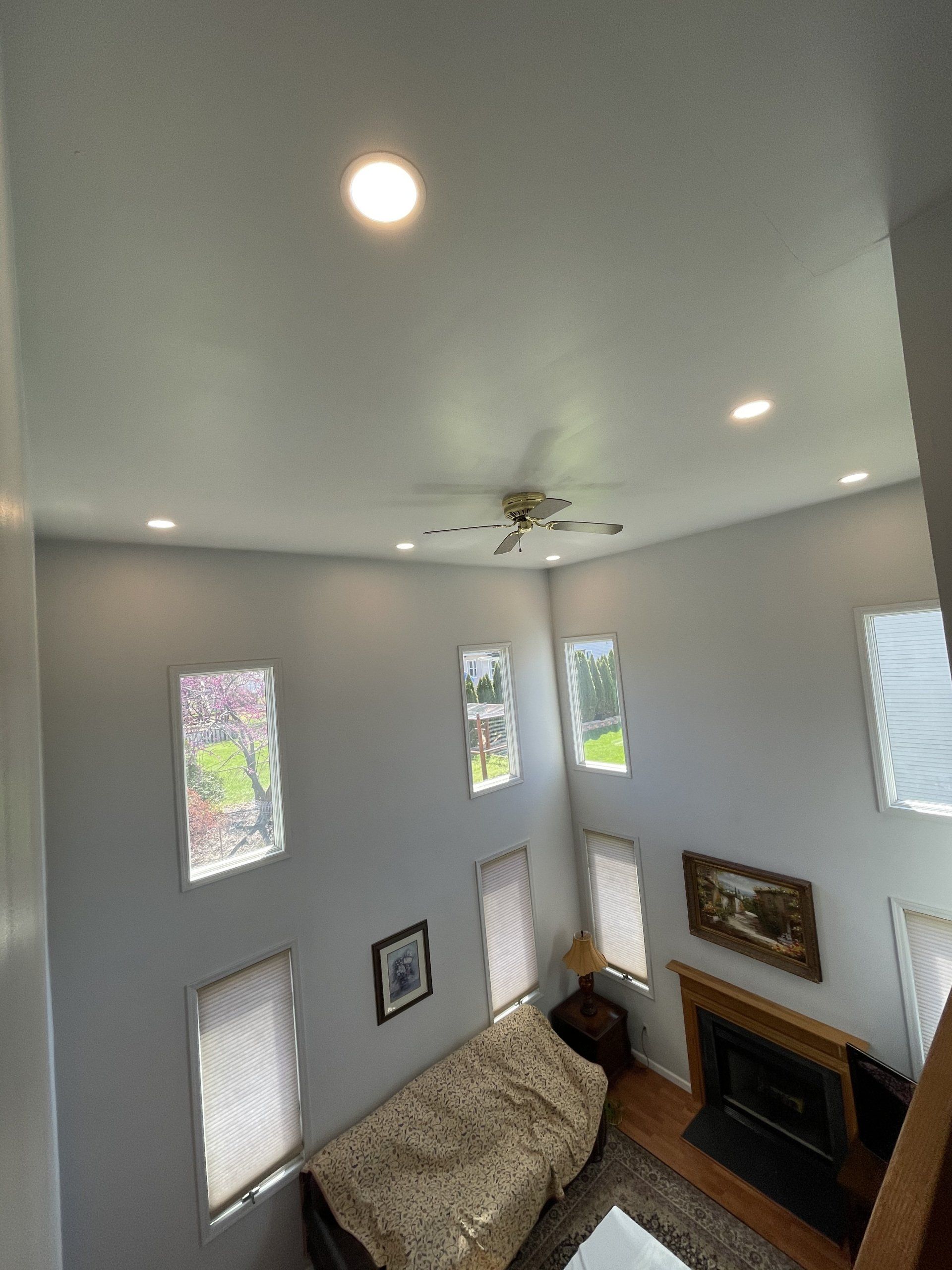 High-ceilinged living room with multiple windows, recessed lights, and a ceiling fan; a sofa and fireplace are visible.