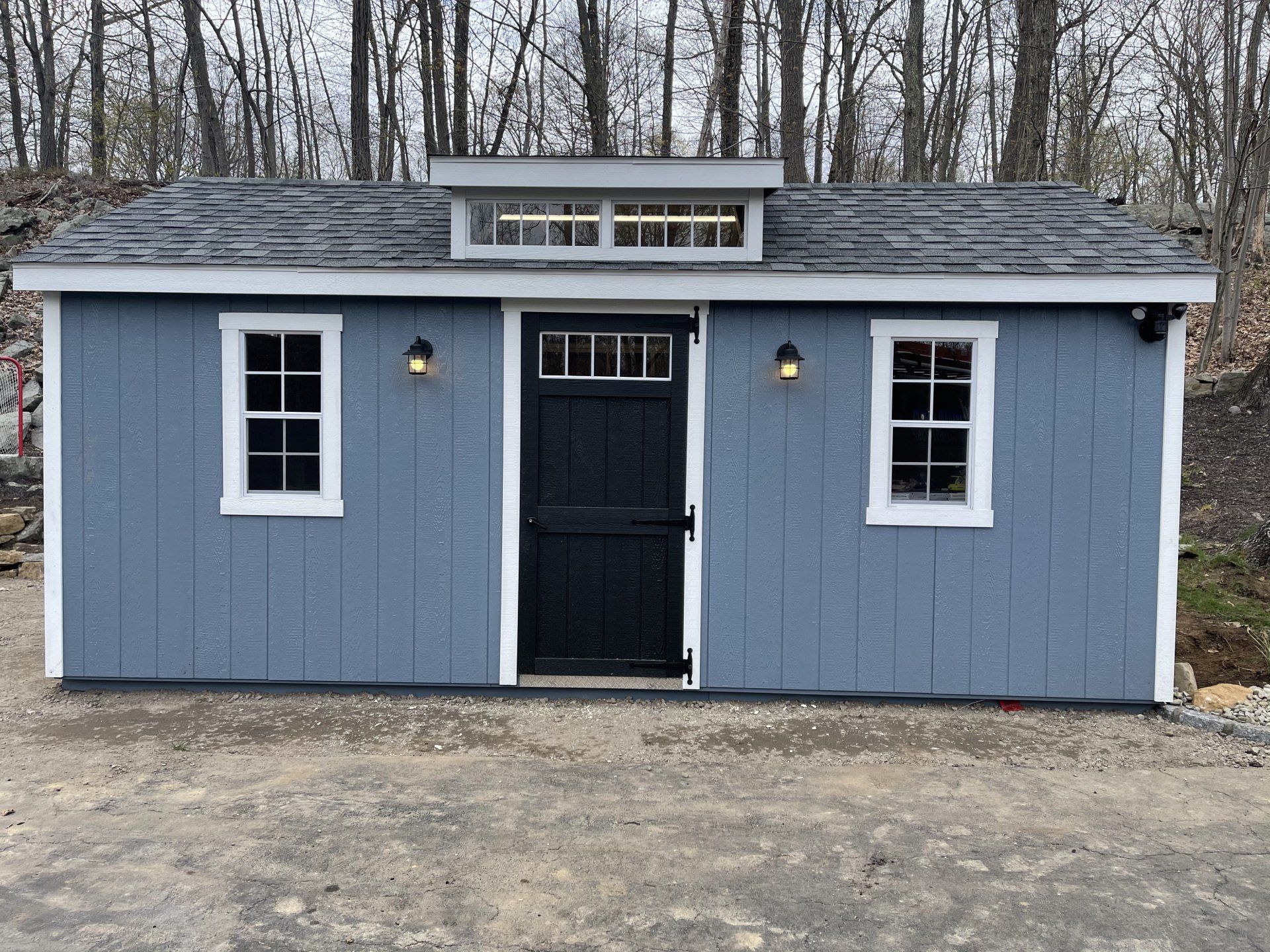 Blue shed with white-trimmed windows and door, black door, and small upper window.