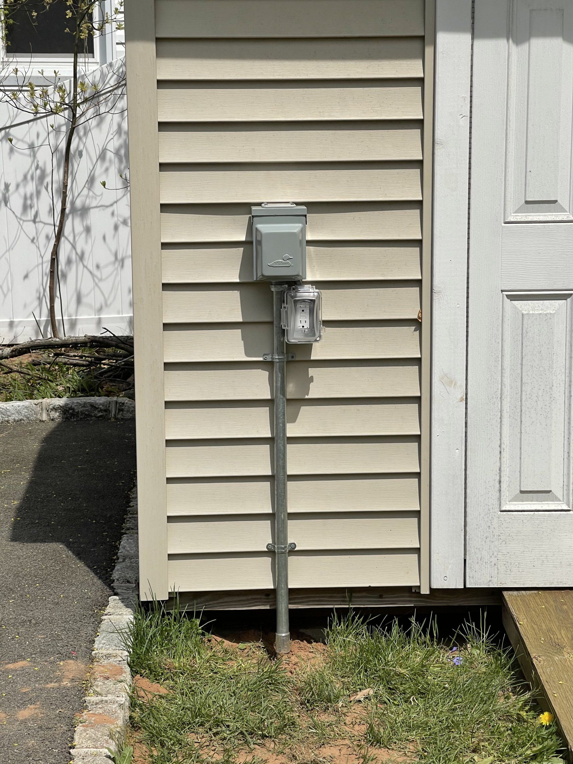 Electrical box and light on siding next to white door. Green grass and asphalt below.