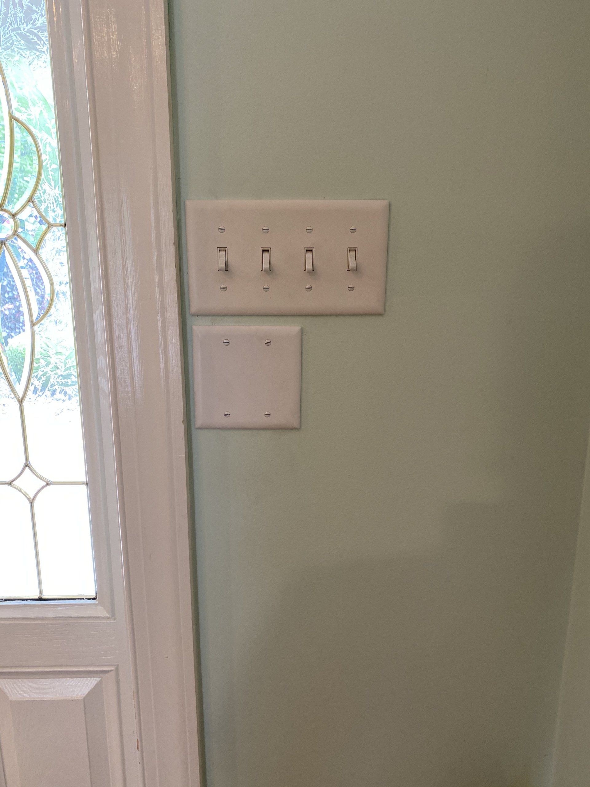 Light switches on a pale green wall, below a blank cover plate, next to a white door with a stained-glass panel.