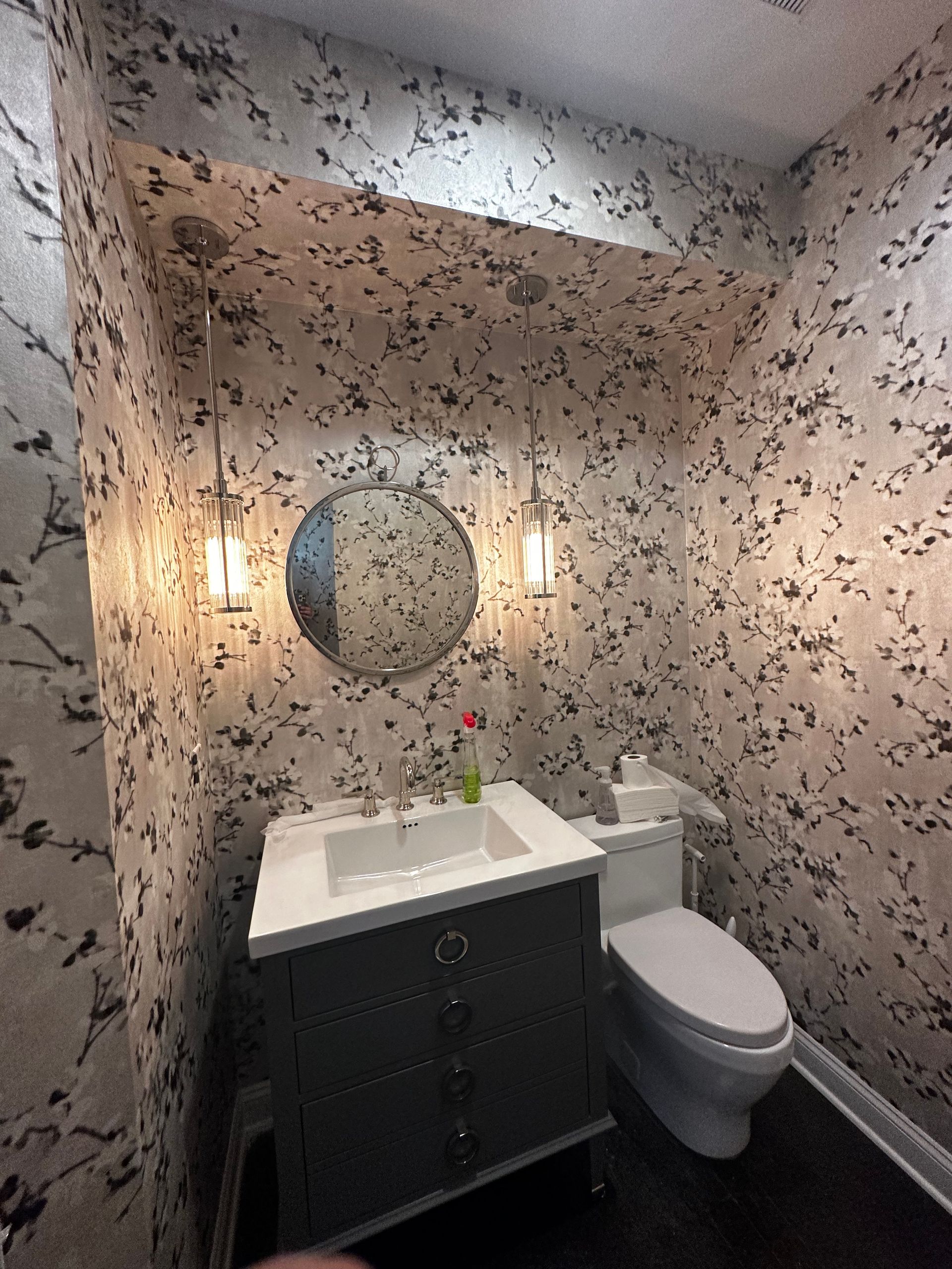 Small bathroom with speckled wallpaper, a gray vanity, round mirror, and a toilet.