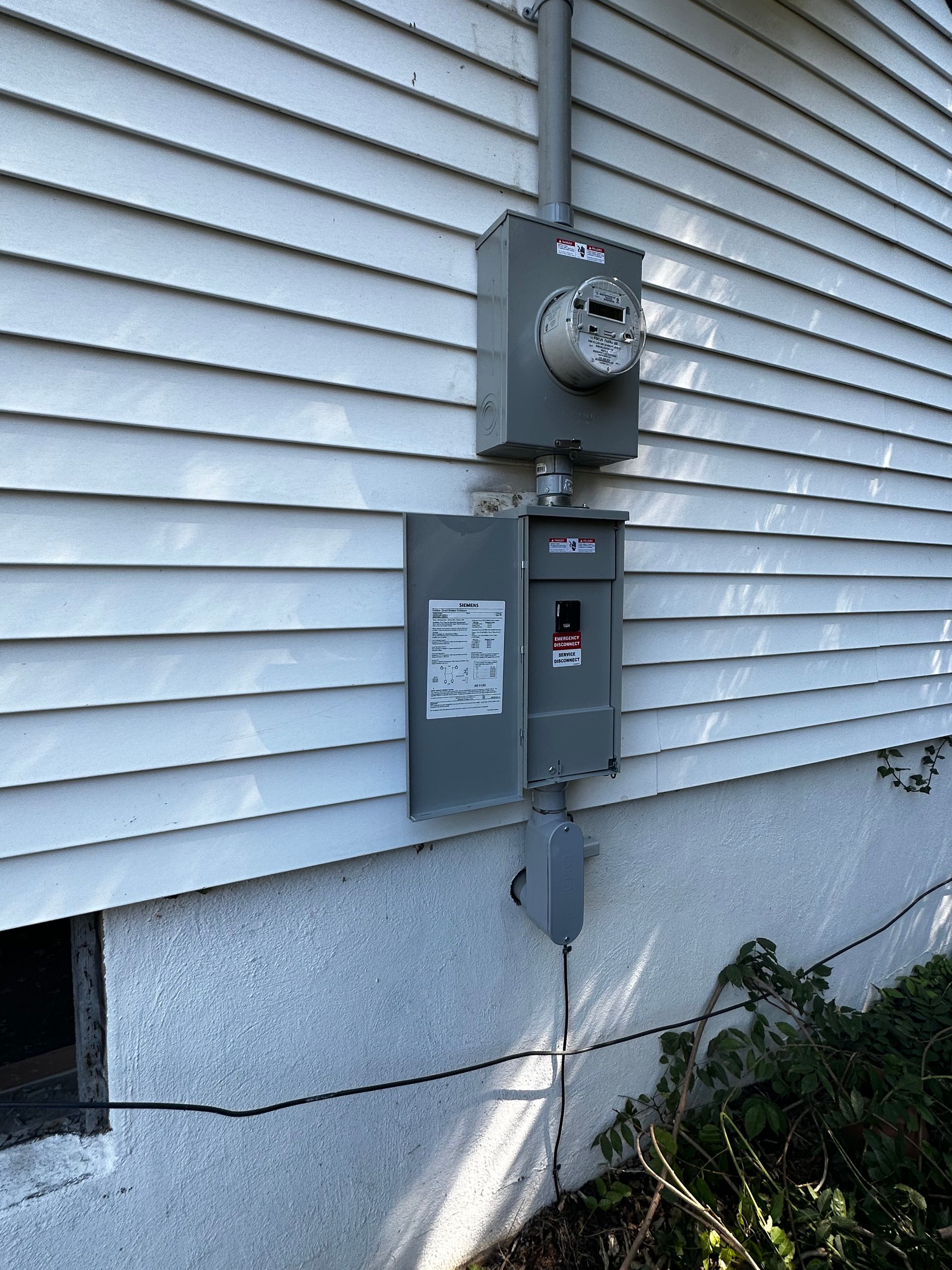 Exterior electrical meter and breaker box mounted on a white clapboard wall.