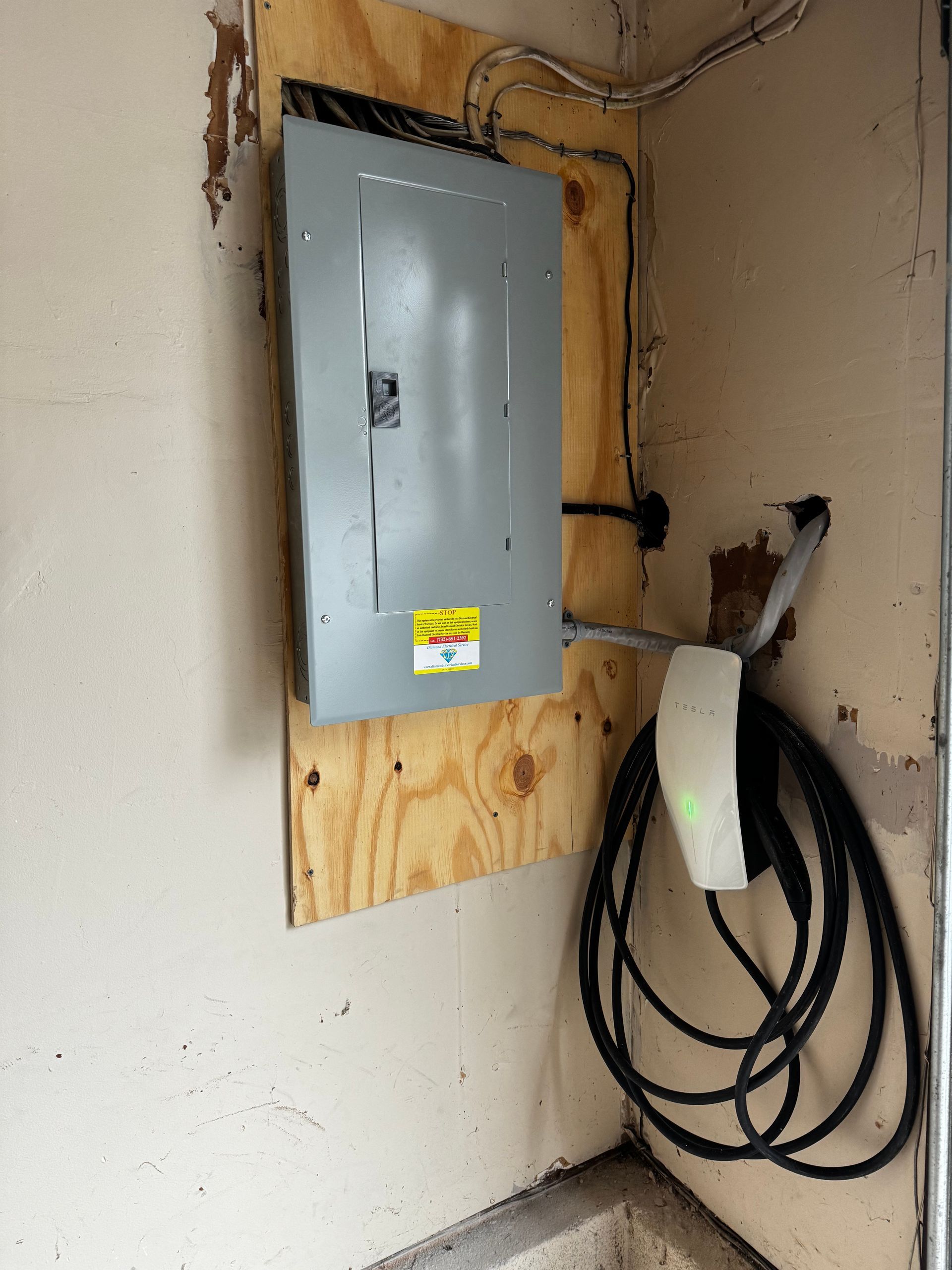 Electrical panel on plywood mounted on a cracked wall, with coiled black cable hanging below.