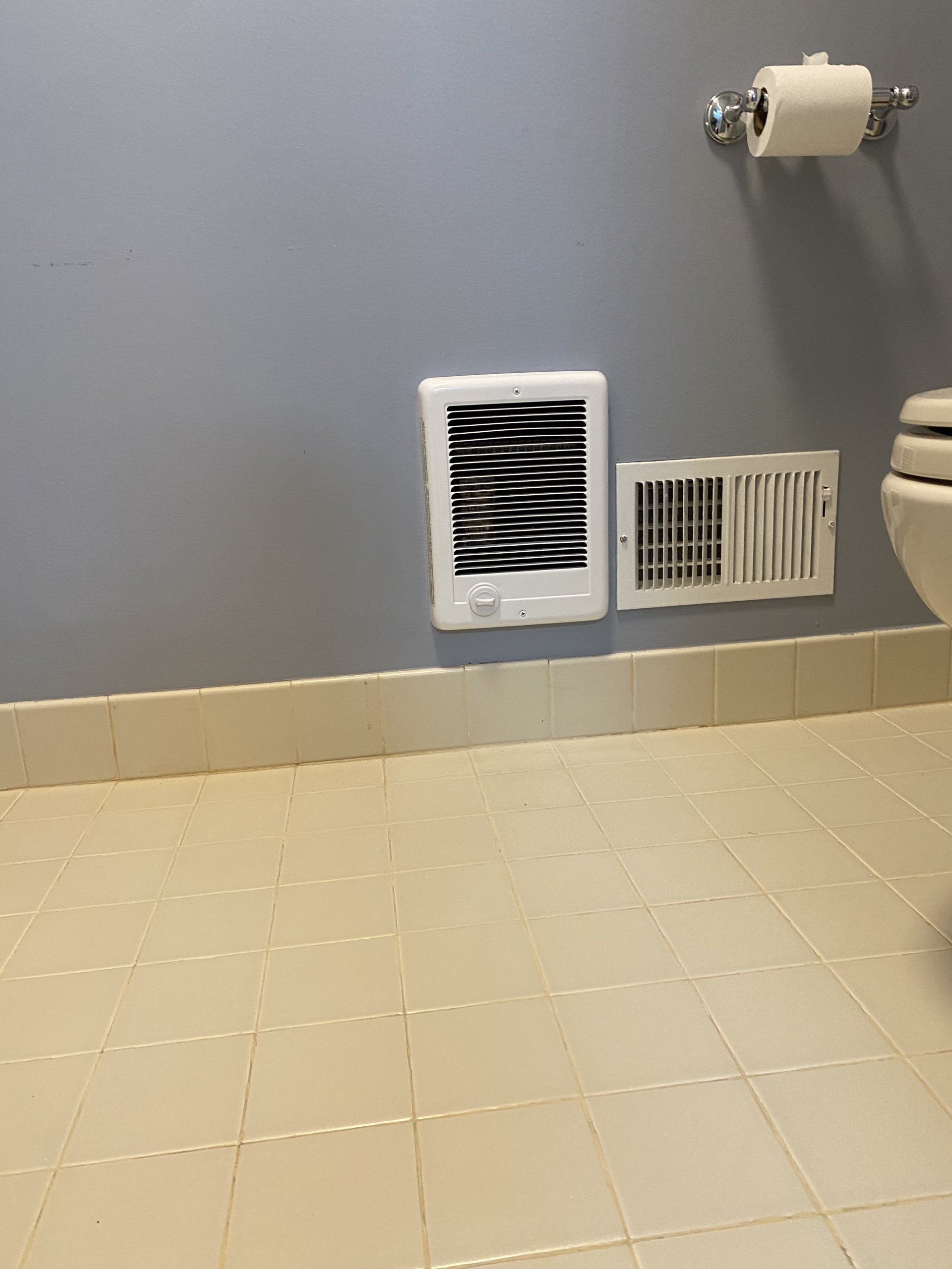 White wall heater and vent on a blue bathroom wall, next to a toilet and a roll of toilet paper.