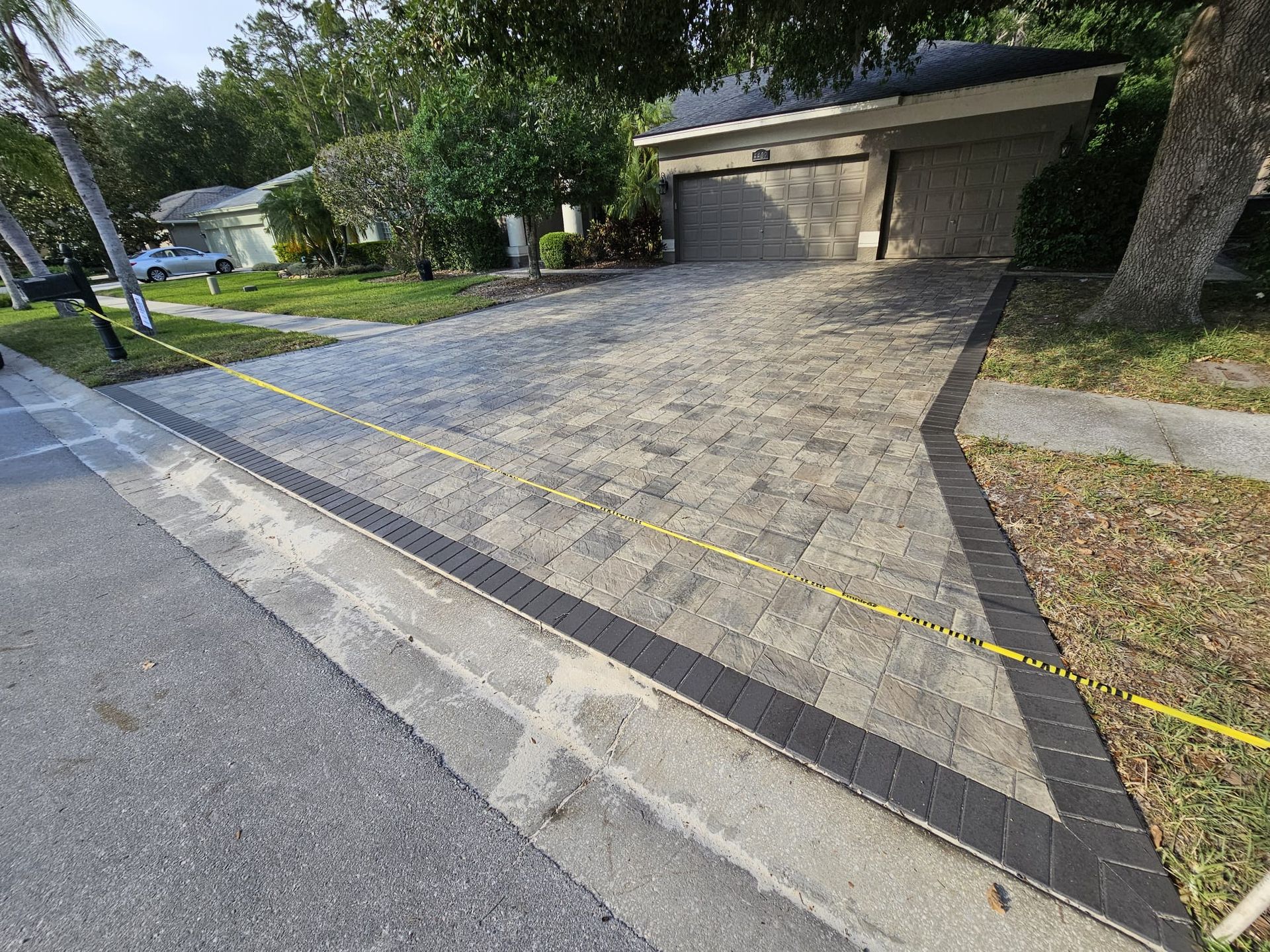 Brick driveway with black border, next to a sidewalk and lawn, taped off with yellow tape.