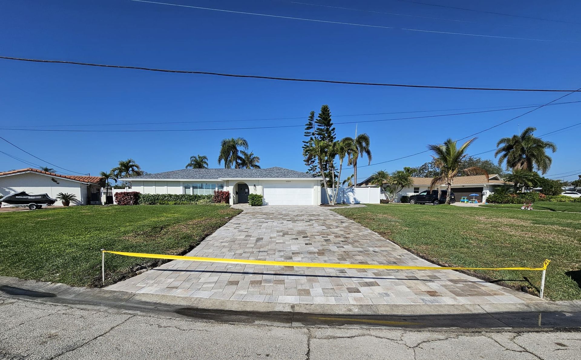 A brick driveway leads to a white house with a garage, blue sky, palm trees. Yellow tape blocks the driveway.