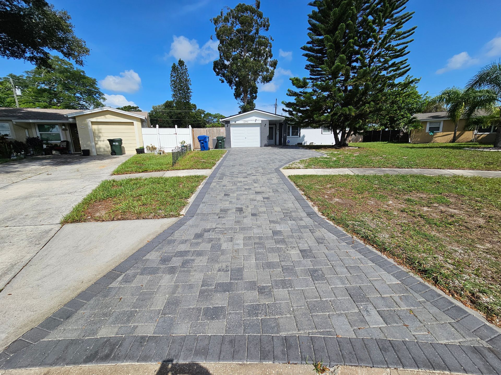 Driveway paved with grey bricks leading to a white house with green lawn and blue sky.