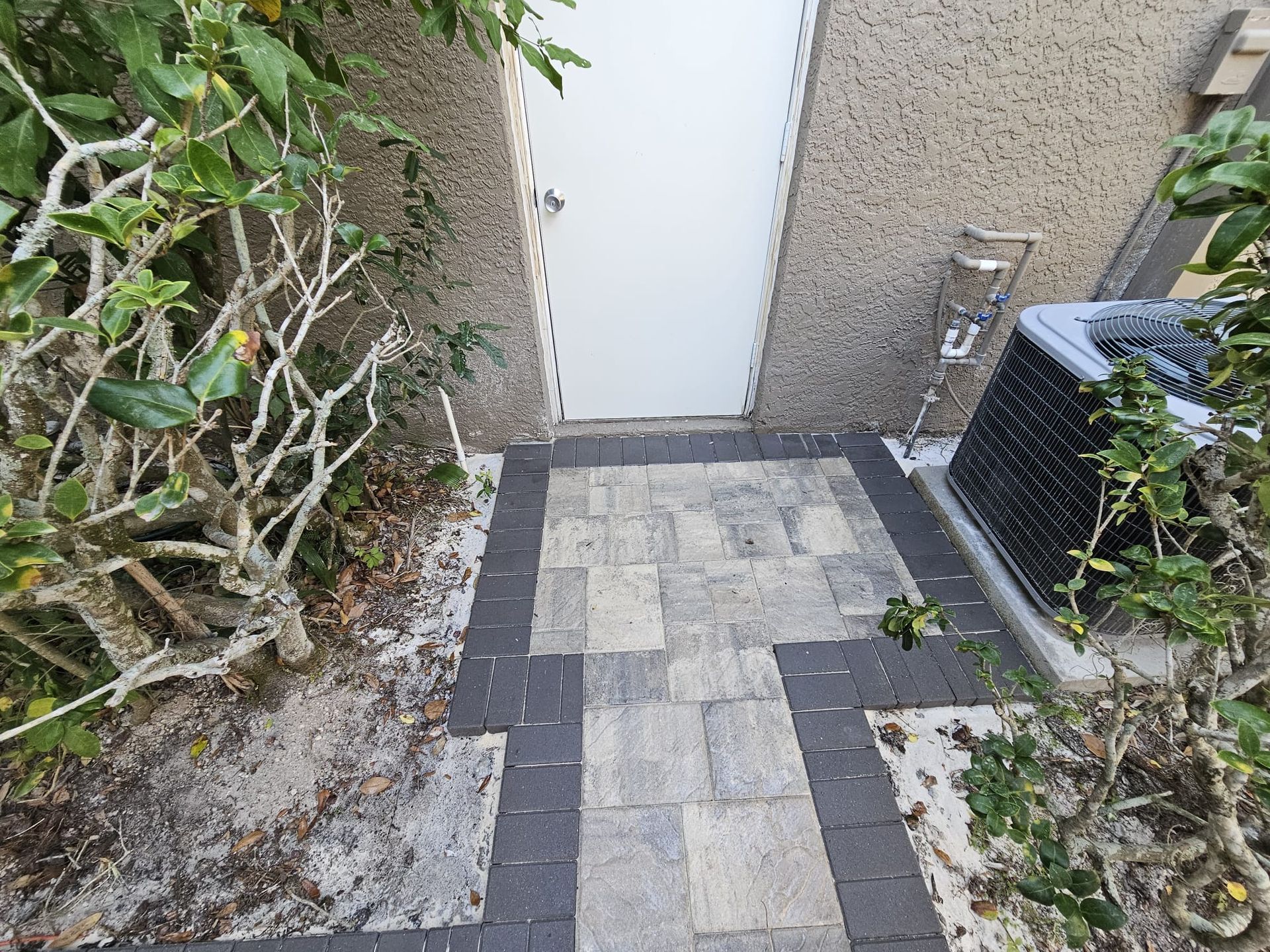 Brick pathway leading to a white door, flanked by shrubs, next to an AC unit.