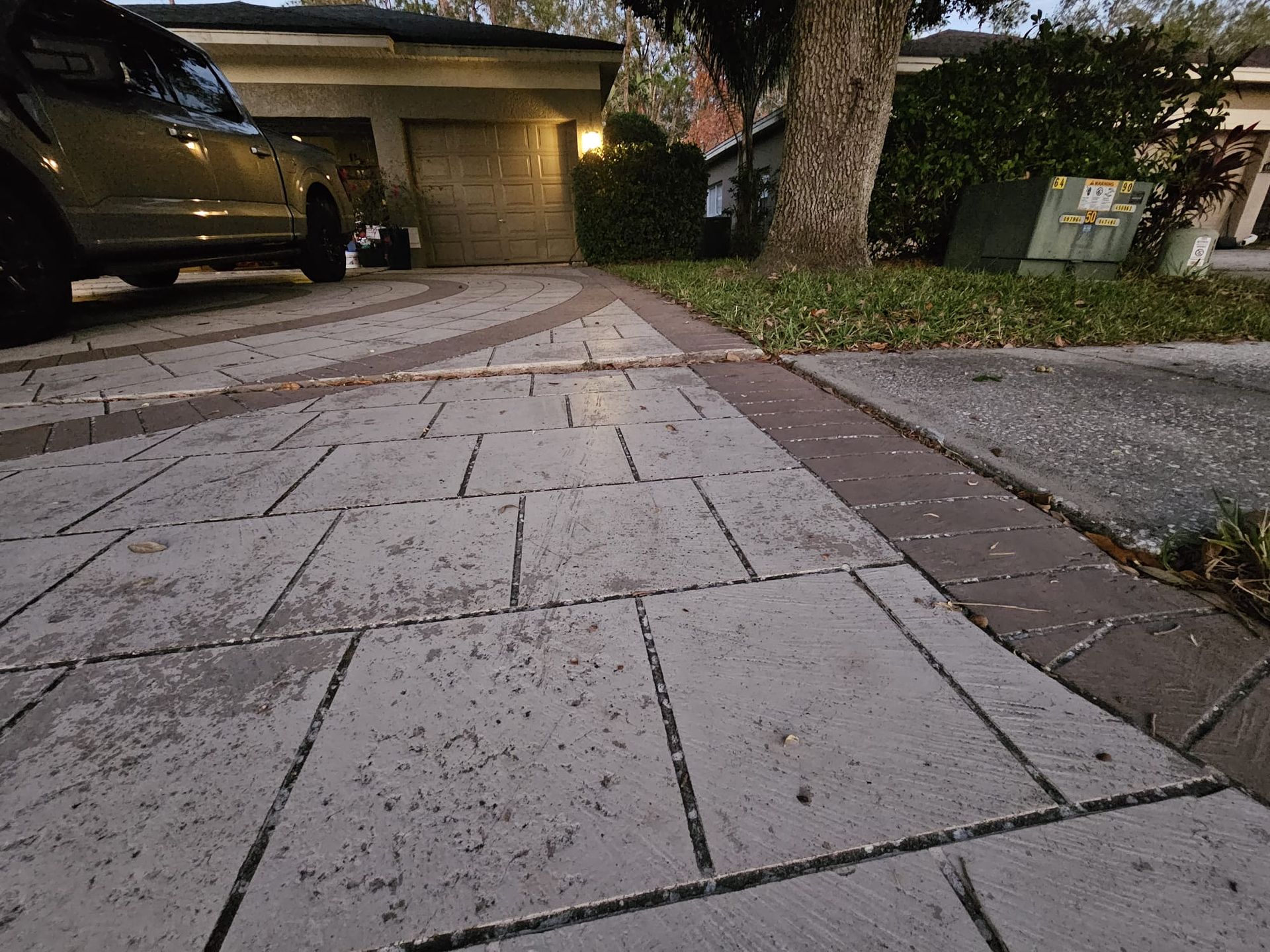 A gray brick driveway leads to a garage, a car parked nearby; trees and grass border the edge.