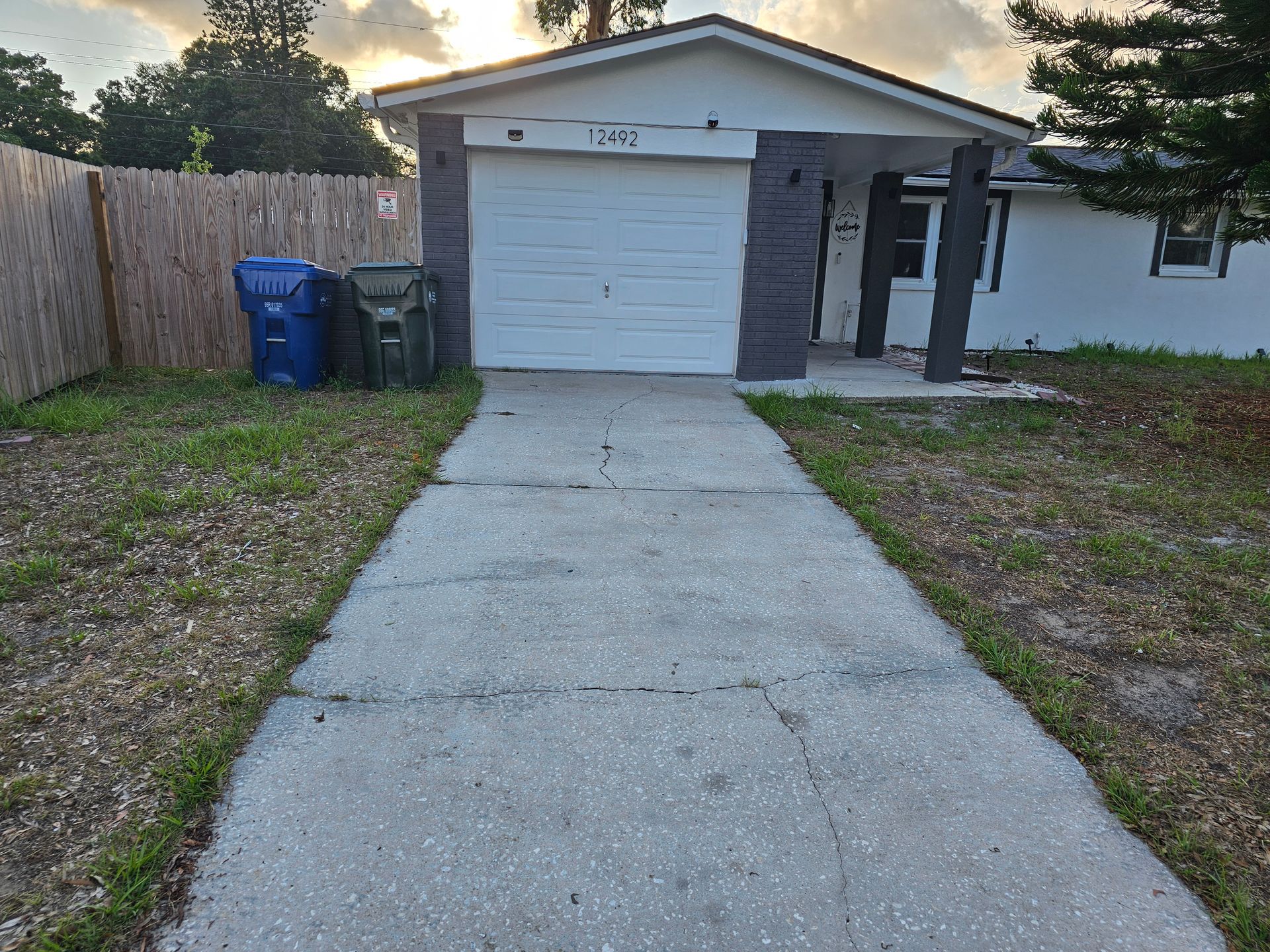 House with gray driveway, garage, and entrance, with trash cans on left.