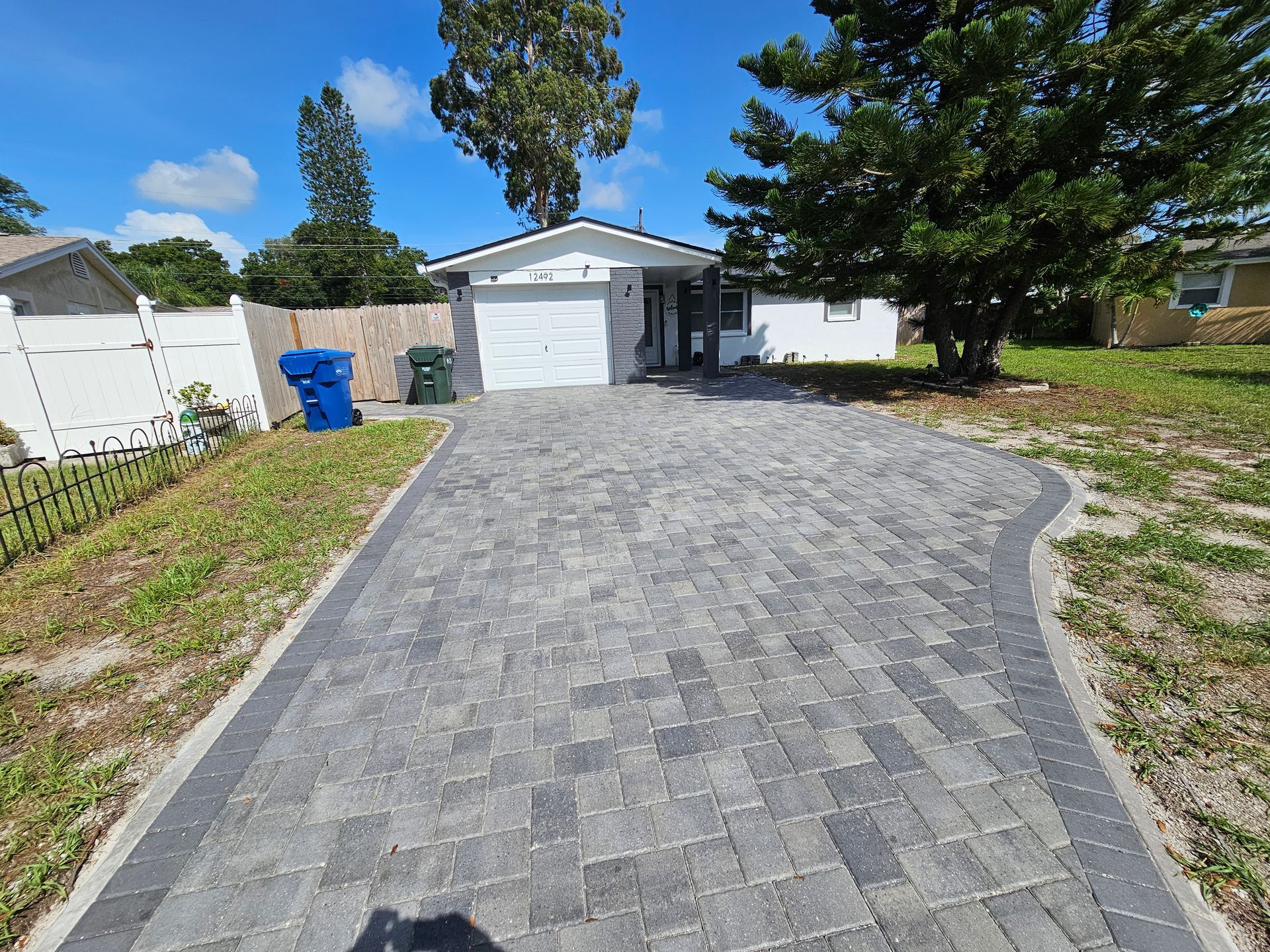 Gray brick paver driveway leading to a white house with a garage door, under a blue sky.