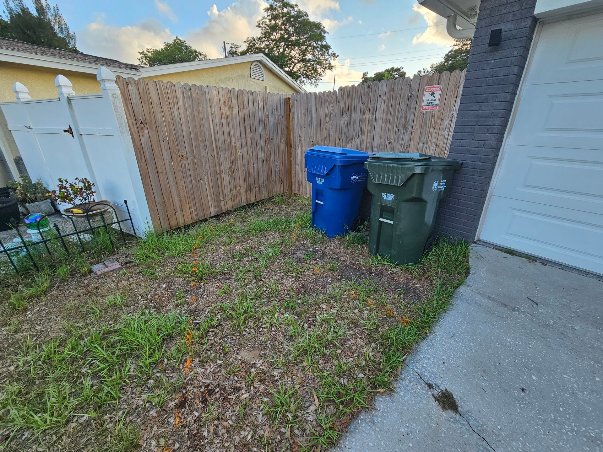 Two trash bins near a wooden fence and a garage; weeds growing in the grassy area.