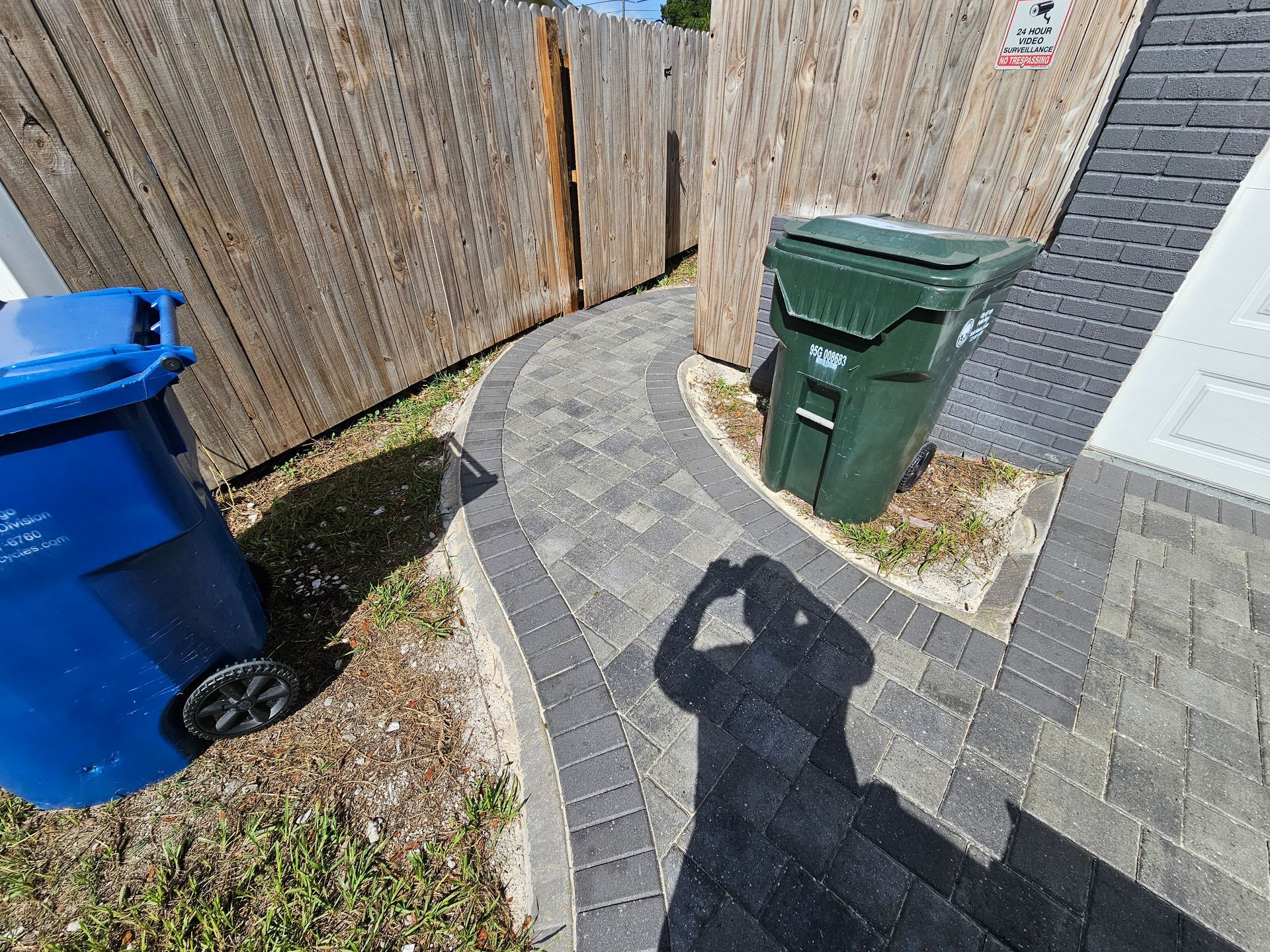 A paved pathway curves past a green and blue trash can in a yard, with a wooden fence and person's shadow.