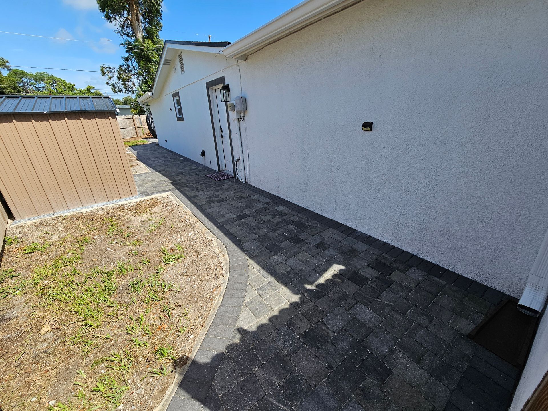 Paved walkway alongside a white stucco building and a small brown shed under a blue sky.