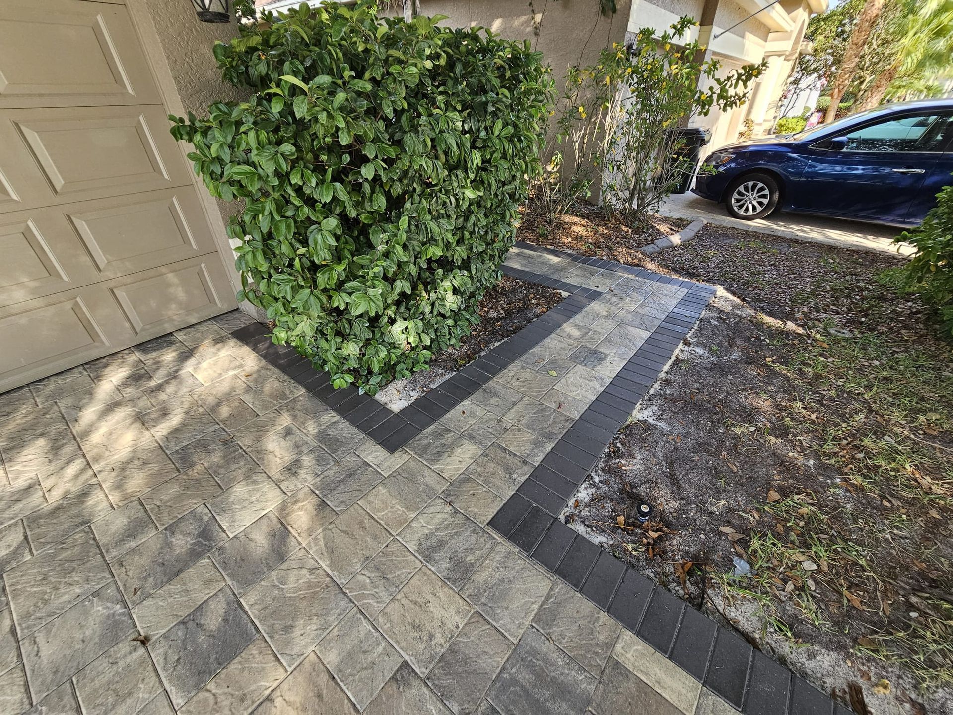 Brick walkway with dark borders, leading to a driveway next to a garage. Lush greenery present.