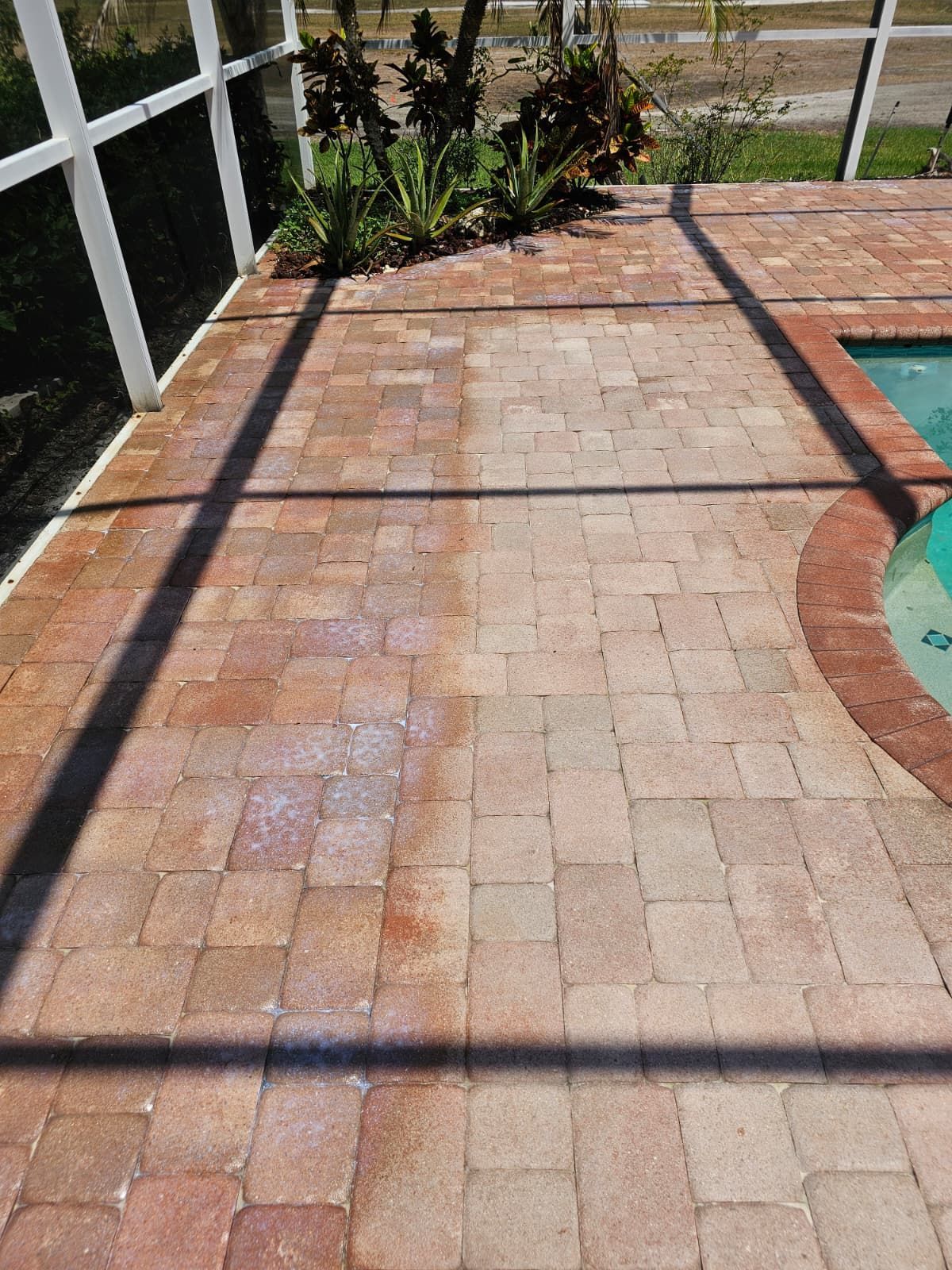 Brick patio, partially cleaned, adjacent to a pool and screened enclosure, plants in the background.