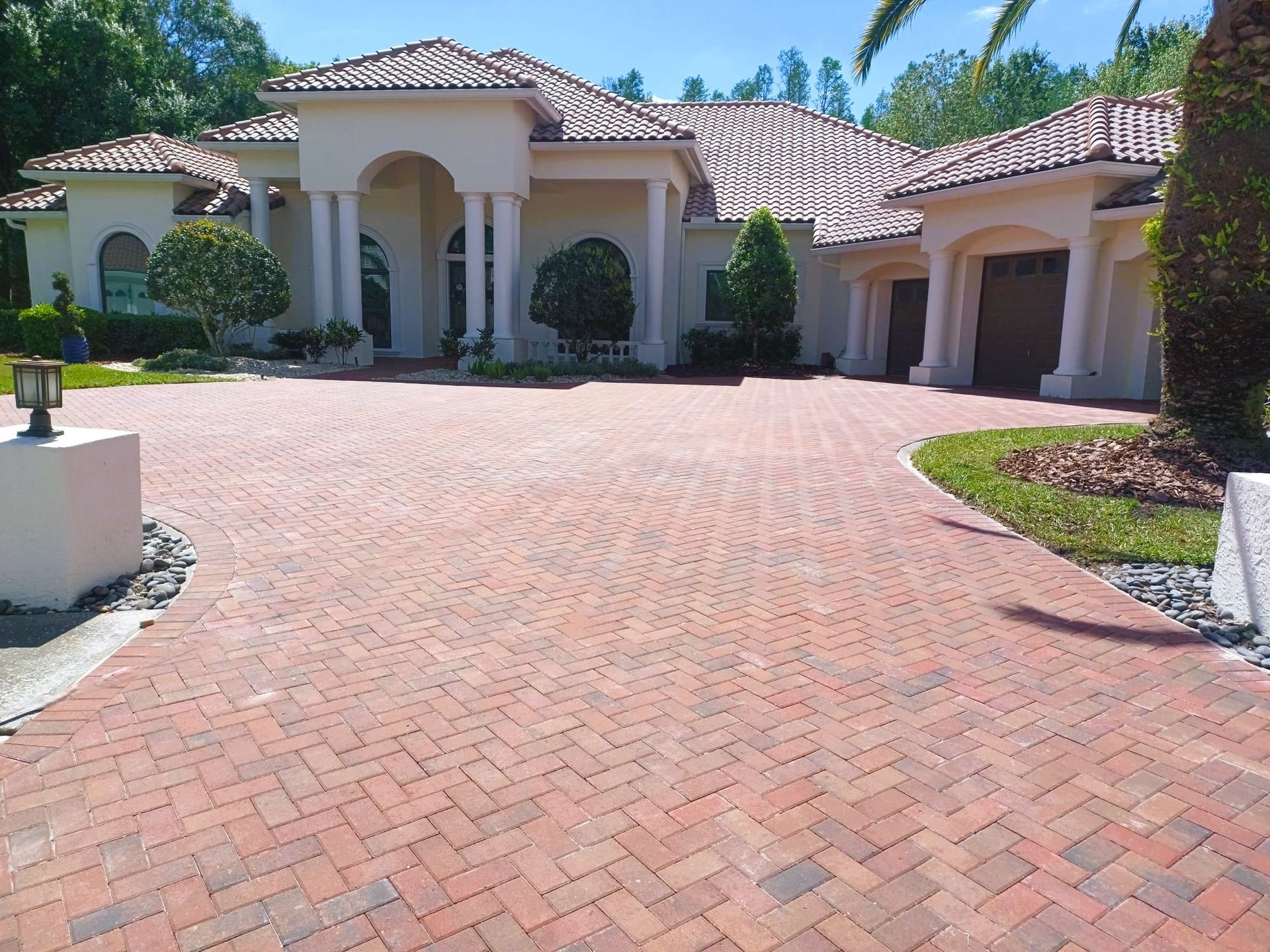 A large beige house with red brick driveway, blue sky, and green trees.