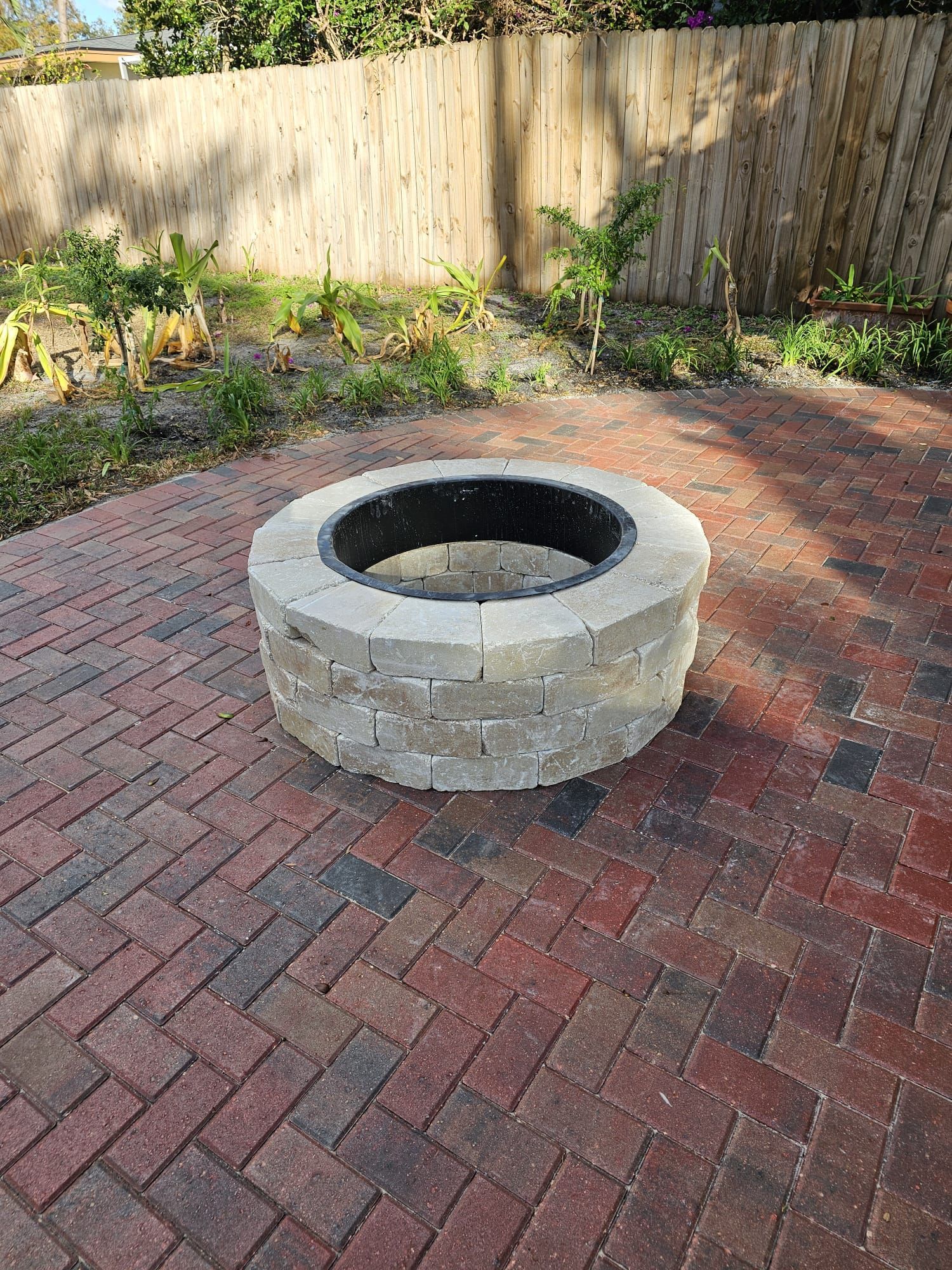 Fire pit made of gray blocks on a brick patio, in a backyard.