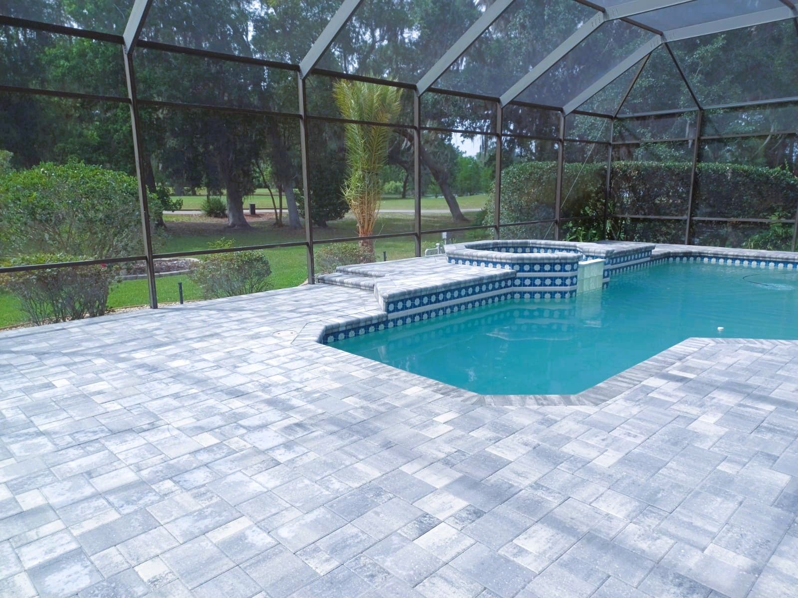 Swimming pool surrounded by blue-tiled deck with spa, enclosed by screen, overlooking a yard.