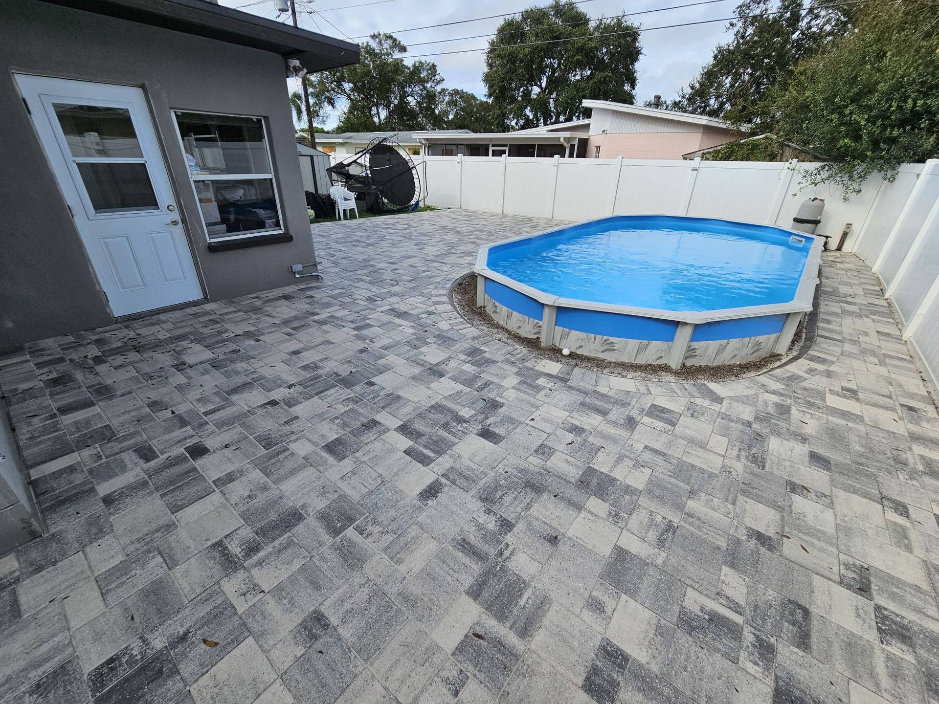 Backyard with an above-ground pool surrounded by gray pavers, near a white fence and building.
