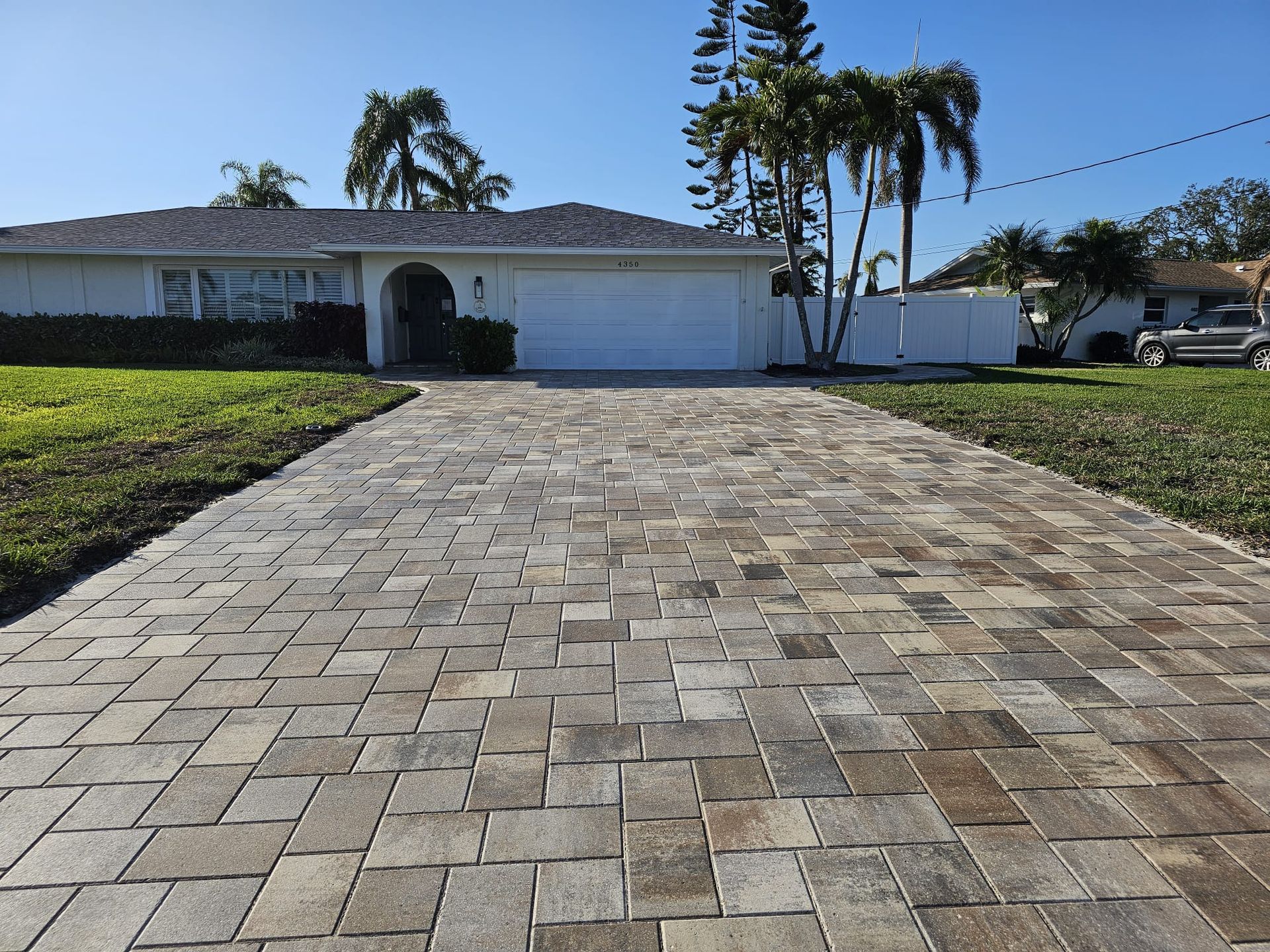 Paver driveway leading to a white house with green grass, palms, and a clear blue sky.