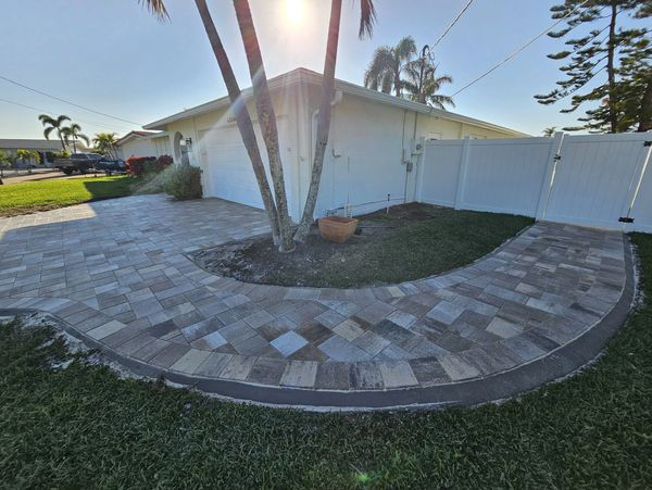 A paved path curves around a tree in front of a white house with a white fence.