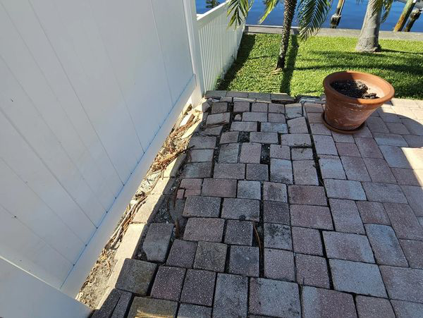 Brick patio next to a white fence, with uneven bricks and a potted plant.