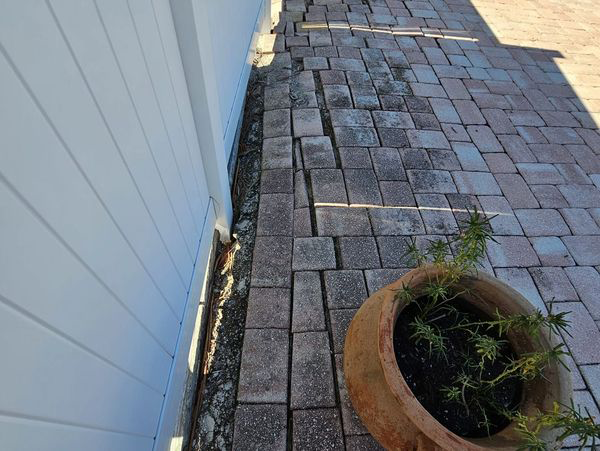 Brick pavers on a walkway are cracked and uneven next to a white wall. A potted plant sits nearby.