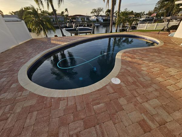 A kidney-shaped pool with a pool cleaner in it, surrounded by red brick and a waterfront view.
