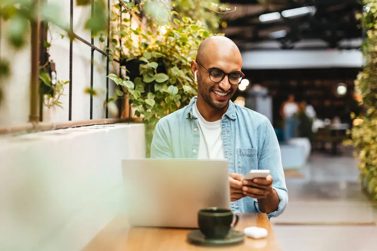 Man sitting outside at a table with his laptop smiling at is phone.