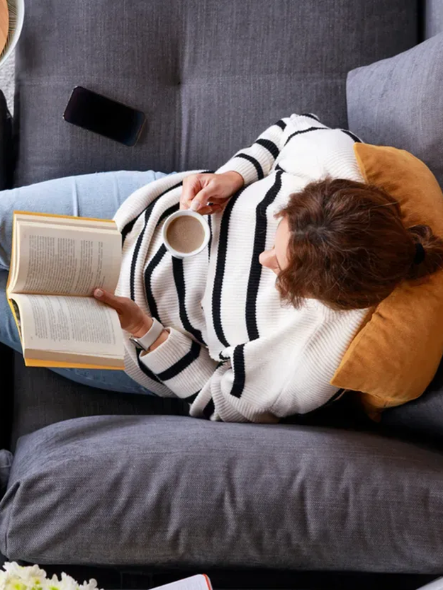 Top-down view of a woman reading on couch drinking coffee.