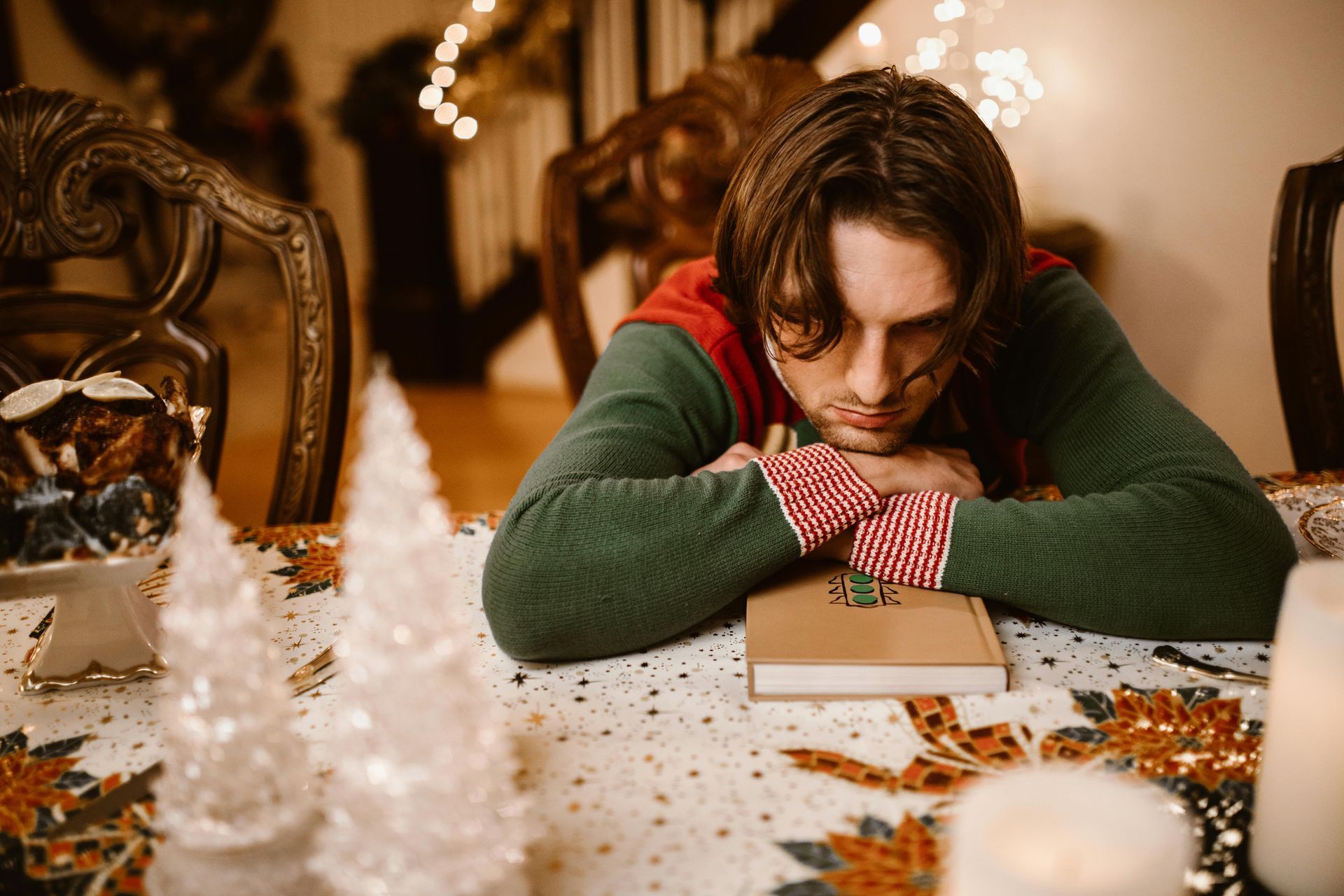 Person in elf attire rests head on table with a book, surrounded by Christmas decorations.