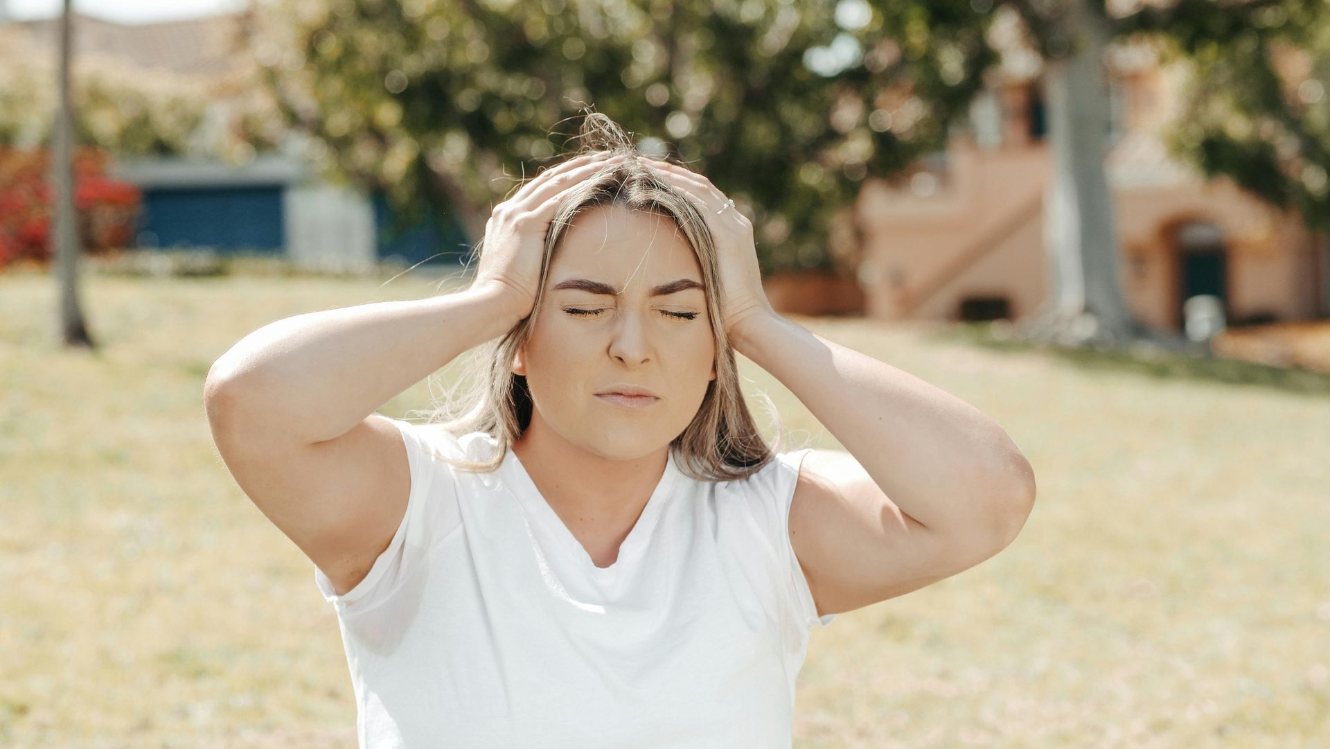 Woman with eyes closed, hands on head, in a park, looking stressed.