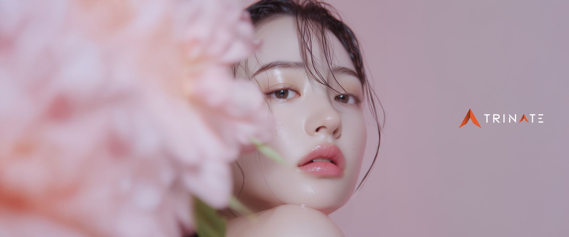 A woman's face peeks from behind pink flowers. She has light skin and her lips are glossy.