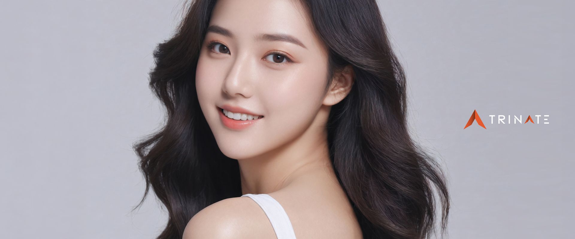 Young woman with long dark wavy hair smiling, wearing a white top, against a grey background.