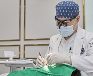 Dentist wearing a surgical mask and cap, working on a patient in a medical setting.