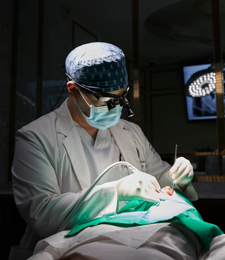 Dentist in white coat with mask, glasses, and headlight operating on a patient's mouth in a well-lit dental office.