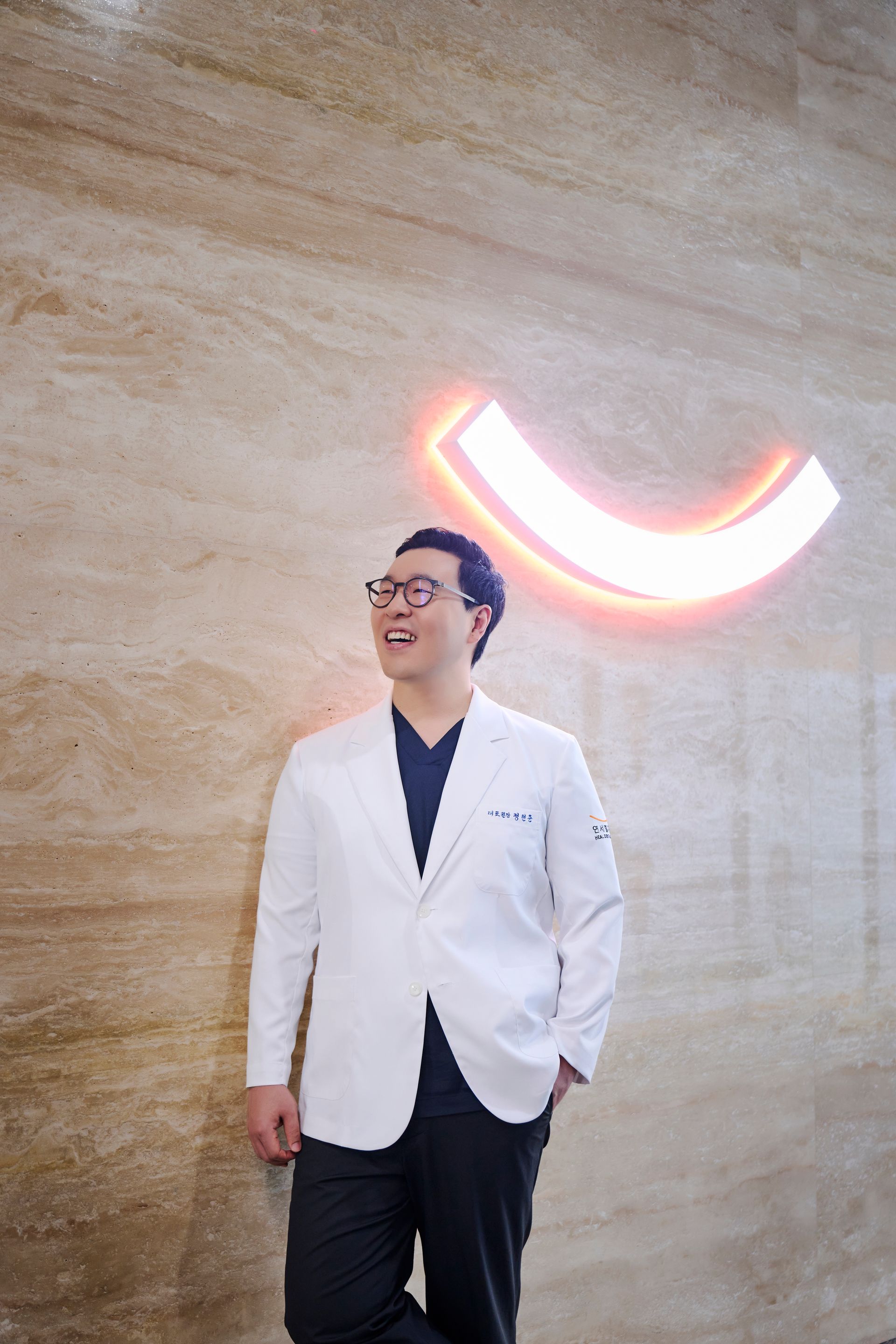 Man in white coat smiles, leaning against a textured wall with a neon-lit curved shape.
