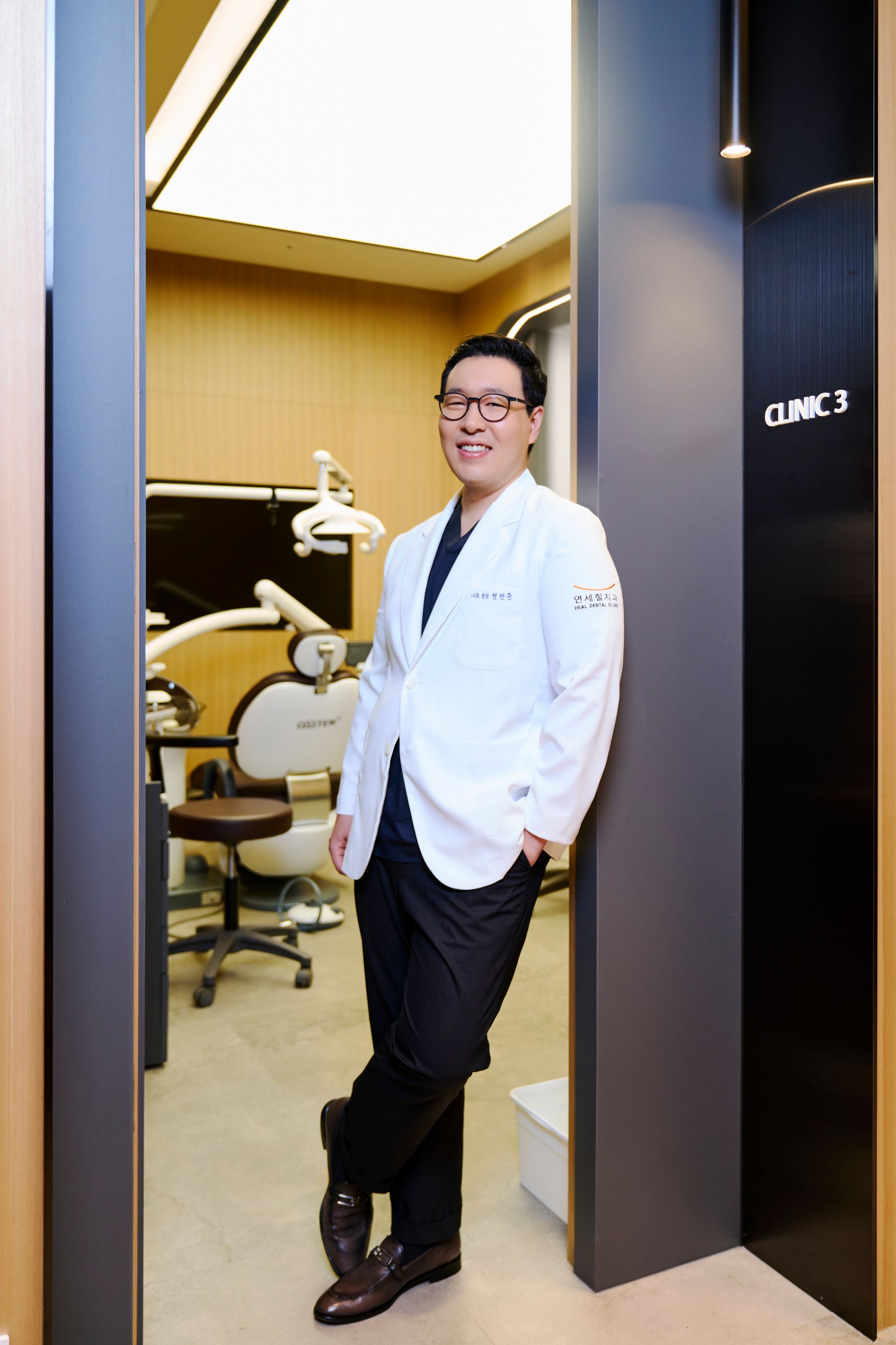 Smiling dentist in a white coat stands in doorway to clinic, leaning on wall.